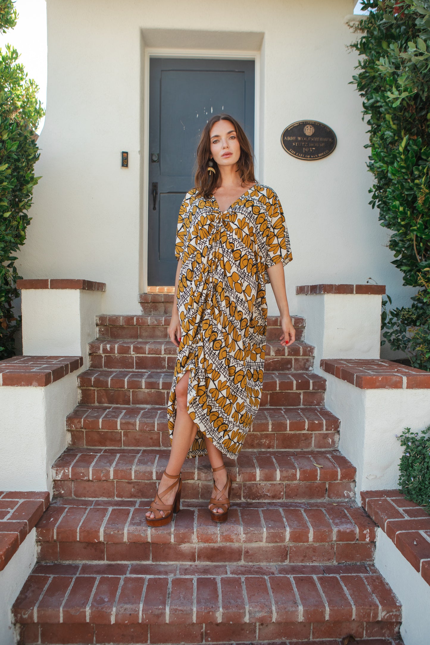 Model wears caftan with beige, taupe and black abstract, mid-century modern, tropical print. The caftan has drawstrings tied at the top of the neck ruching the front of the caftan to desired length.