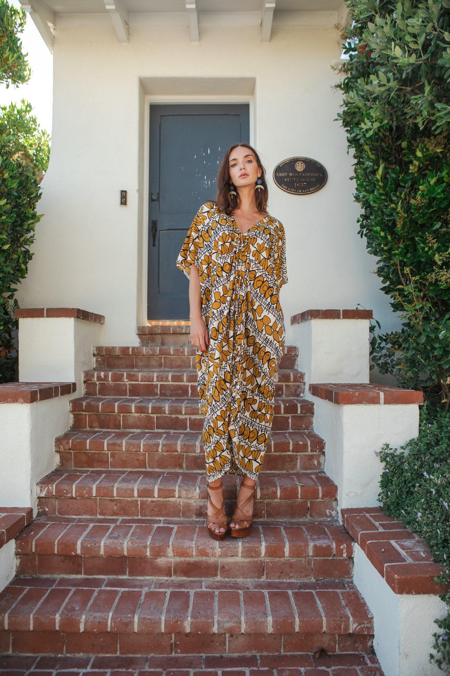 Model wears caftan with beige, taupe and black abstract, mid-century modern, tropical print. The caftan has drawstrings tied at the top of the neck ruching the front of the caftan to desired length.