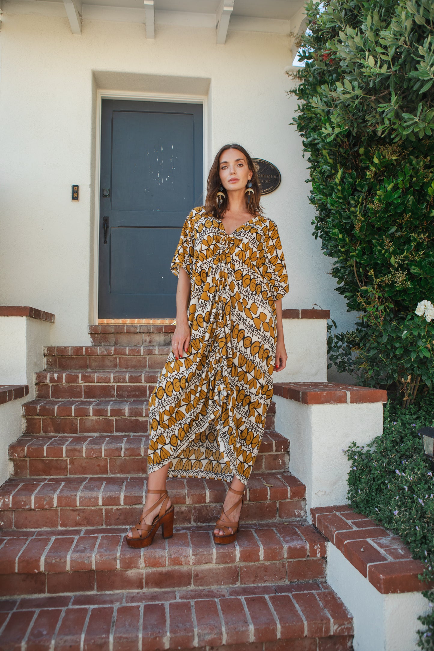 Model wears caftan with beige, taupe and black abstract, mid-century modern, tropical print. The caftan has drawstrings tied at the top of the neck ruching the front of the caftan to desired length.