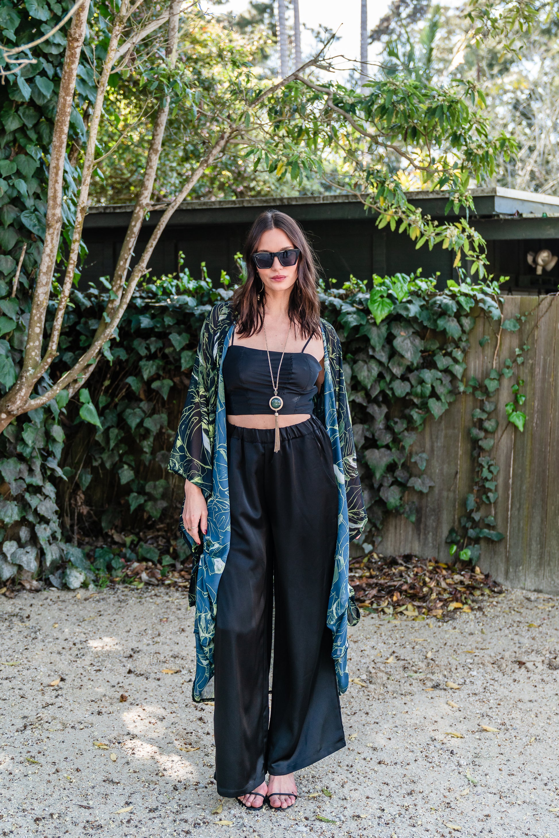 Model wears black chiffon Kimono with varying green palm leaf print. The kimono has classic wide kimono sleeves and lands at midi length. Kimono has a matching tie belt.