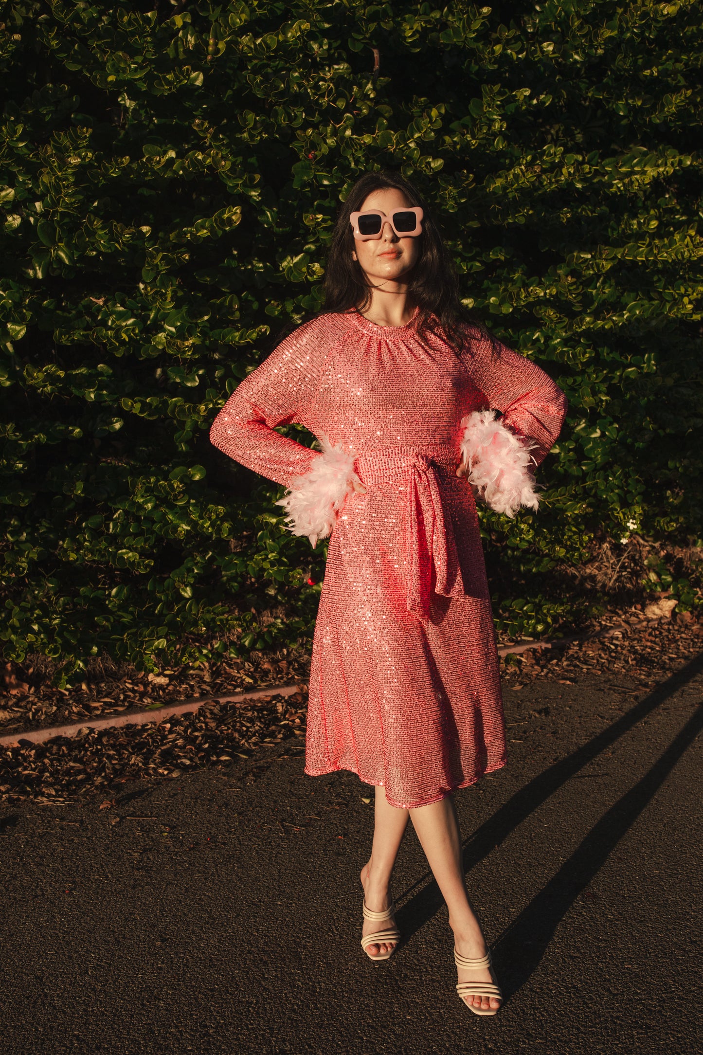 Jennafer Grace light pink flamingo sequins dress. Featuring a boat neck, generous bishop sleeves with light pink feather detail at sleeve hem, flowing midi length maxi skirt, and a slim matching sash for a cinched waist.