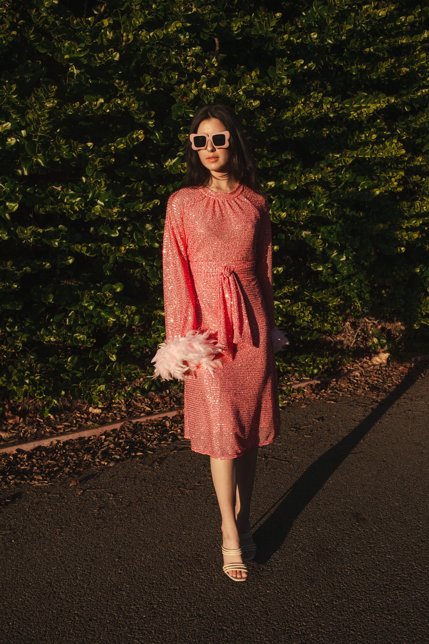 Jennafer Grace light pink flamingo sequins dress. Featuring a boat neck, generous bishop sleeves with light pink feather detail at sleeve hem, flowing midi length maxi skirt, and a slim matching sash for a cinched waist.