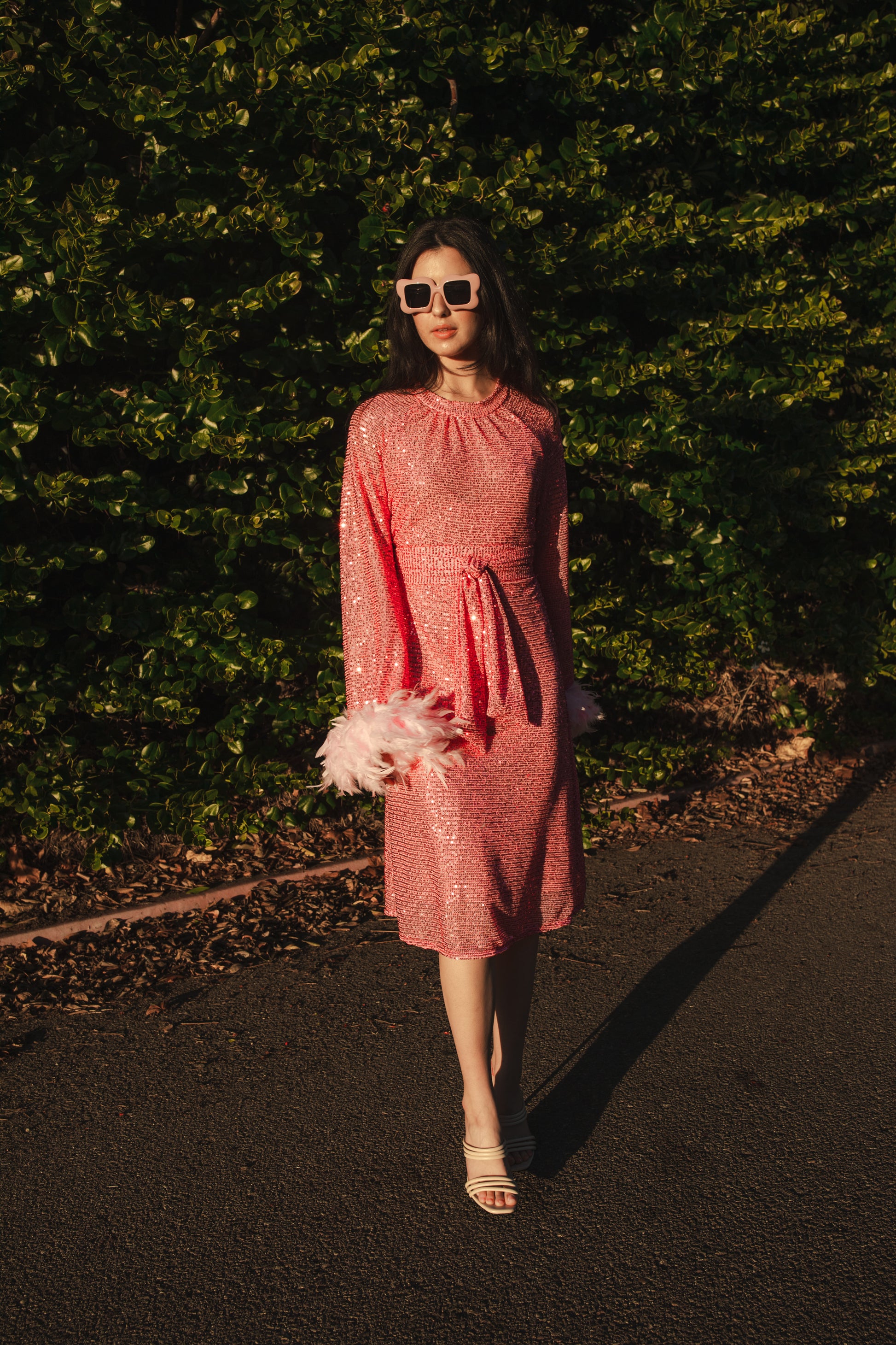 Jennafer Grace light pink flamingo sequins dress. Featuring a boat neck, generous bishop sleeves with light pink feather detail at sleeve hem, flowing midi length maxi skirt, and a slim matching sash for a cinched waist.