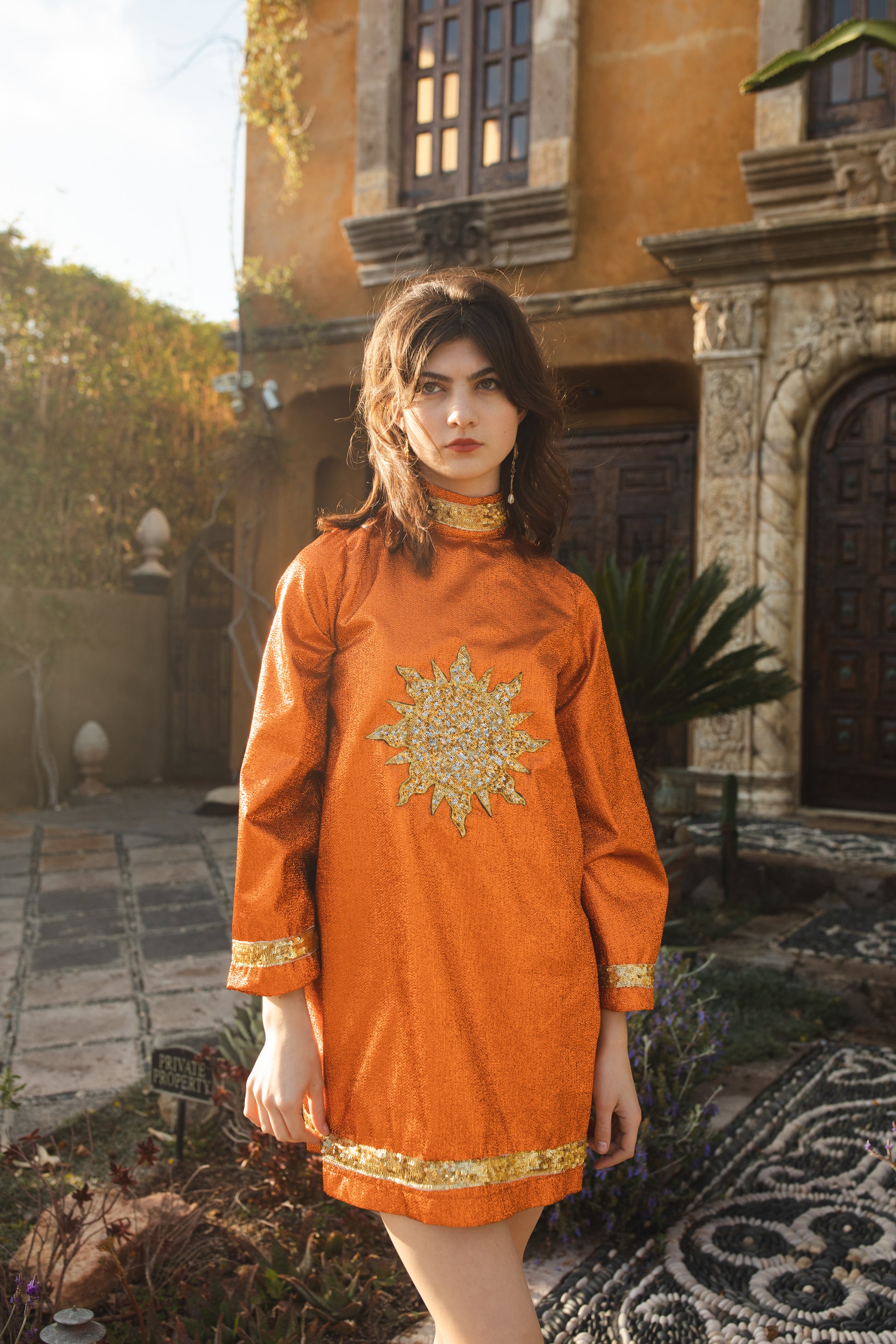 Crafted from a shimmering, fiery orange fabric embellished with an intricate sequin sun and matching sequin details at collar, cuffs, and hem, this is the bold and dreamy 60s revival romance dress every wardrobe craves