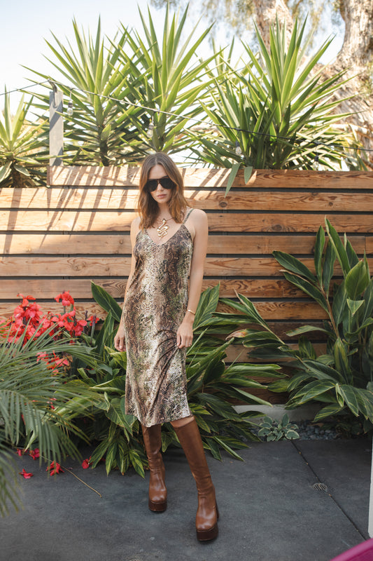 Midi slip dress with a soft beige base color, adorned with snakeskin pattern featuring shades of green and brown. It features slender straps with a straight silhouette and relaxed fix, allowing for a beautiful drape and flowy movement. It's designed for a light, airy feel, suitable for warm weather or layered for cooler days.