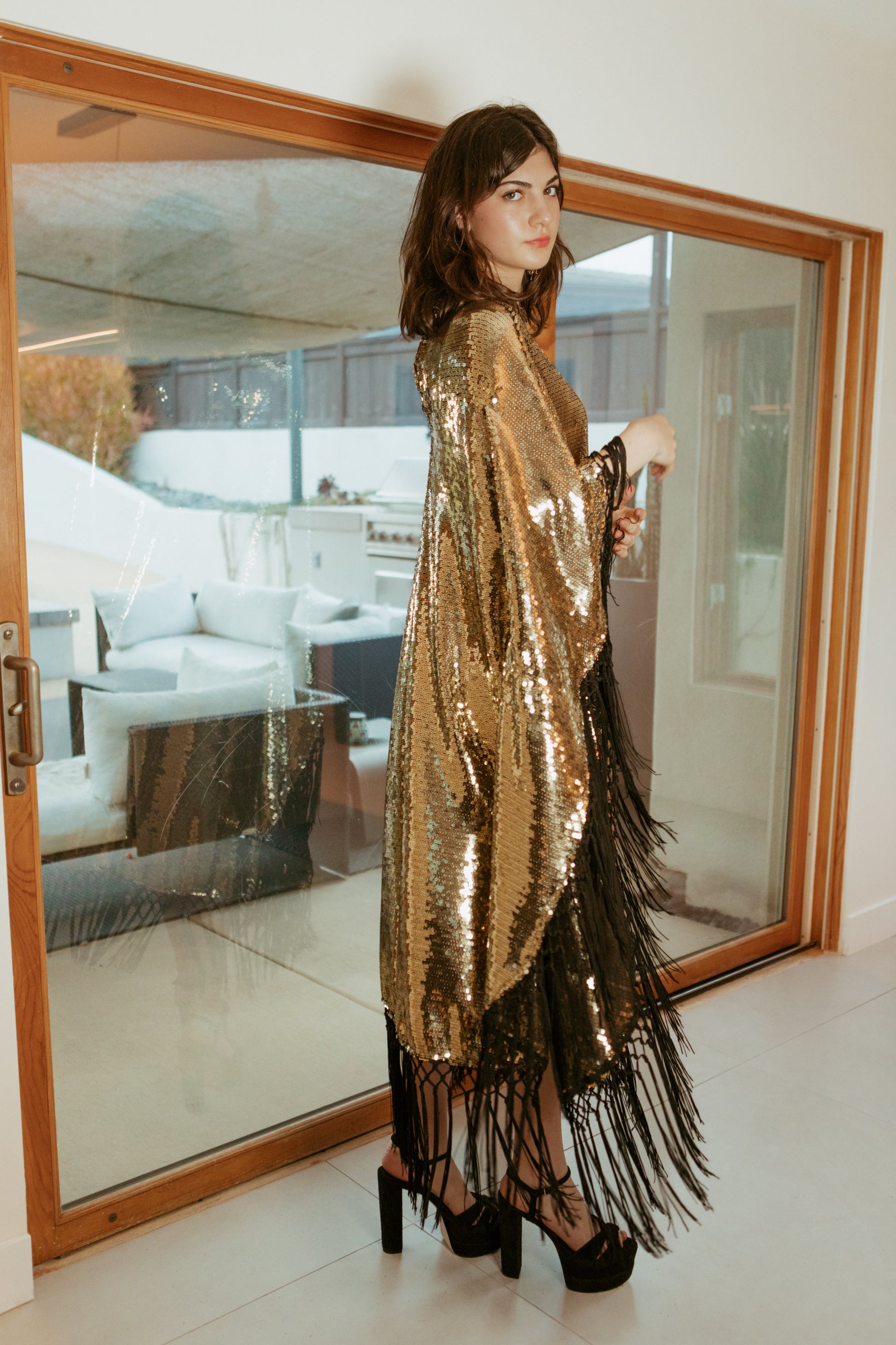 This caftan is a dazzling, full-length garment featuring a shimmering gold sequin overlay on a mesh fabric. The sequins catch the light, creating a sparkling effect throughout. It has a mock neck collar with a straight, loose fit that drapes elegantly over the body. The edges of the caftan, including the hem and the sides, are embellished with long, silk black fringe that add movement and a playful flair to the garment.