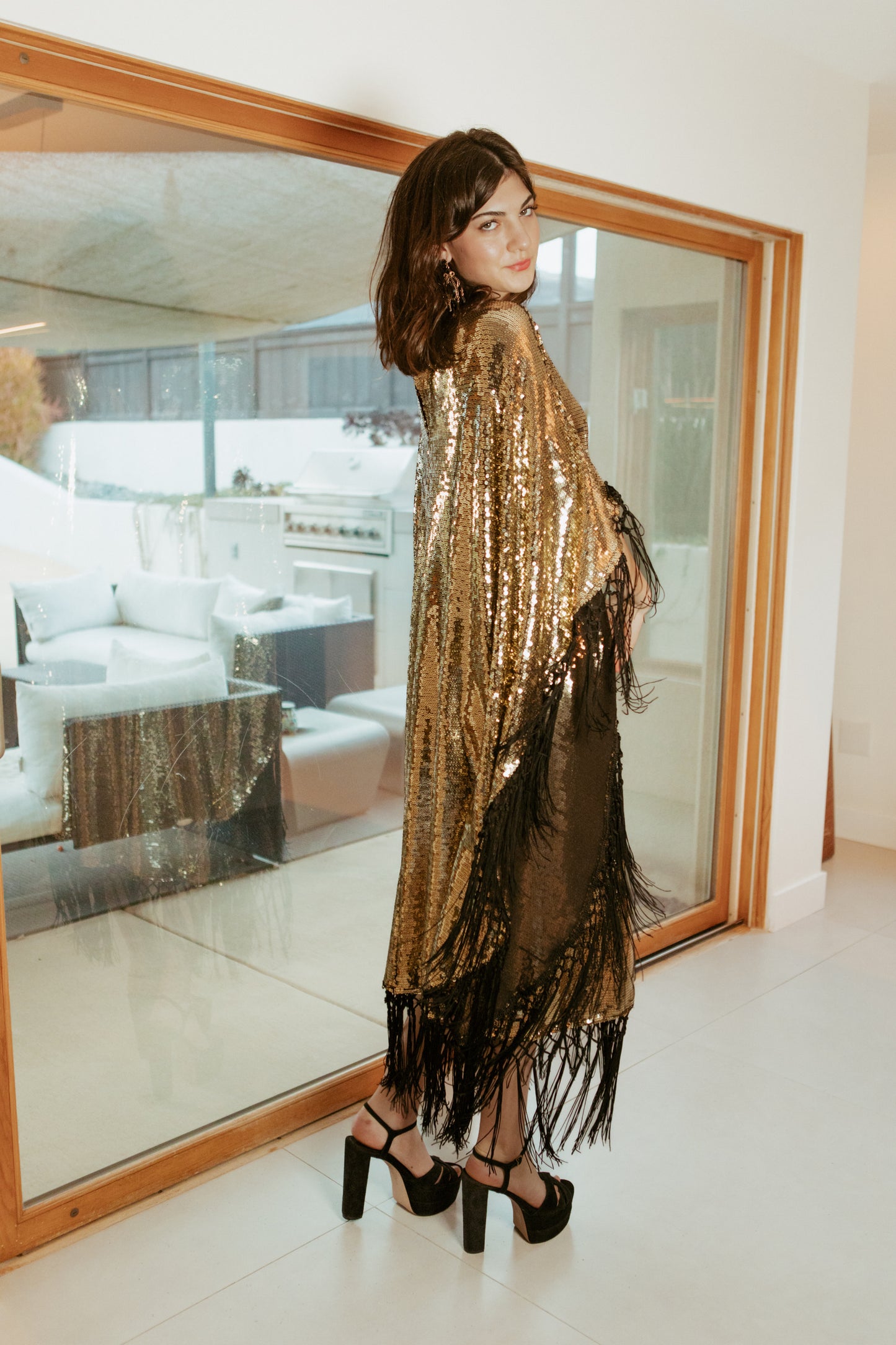 This caftan is a dazzling, full-length garment featuring a shimmering gold sequin overlay on a mesh fabric. The sequins catch the light, creating a sparkling effect throughout. It has a mock neck collar with a straight, loose fit that drapes elegantly over the body. The edges of the caftan, including the hem and the sides, are embellished with long, silk black fringe that add movement and a playful flair to the garment.