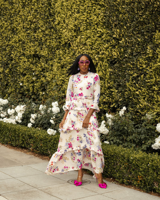 A floral maxi dress made from a soft, linen like crepe, it features high neck, bishop sleeves, tiered ruffle skirt and matching sash for a cinched waist. It's light, airy silhouette. Base color is pale yellow with a mixture of fuchsia, orange, and blue flowers as the print.