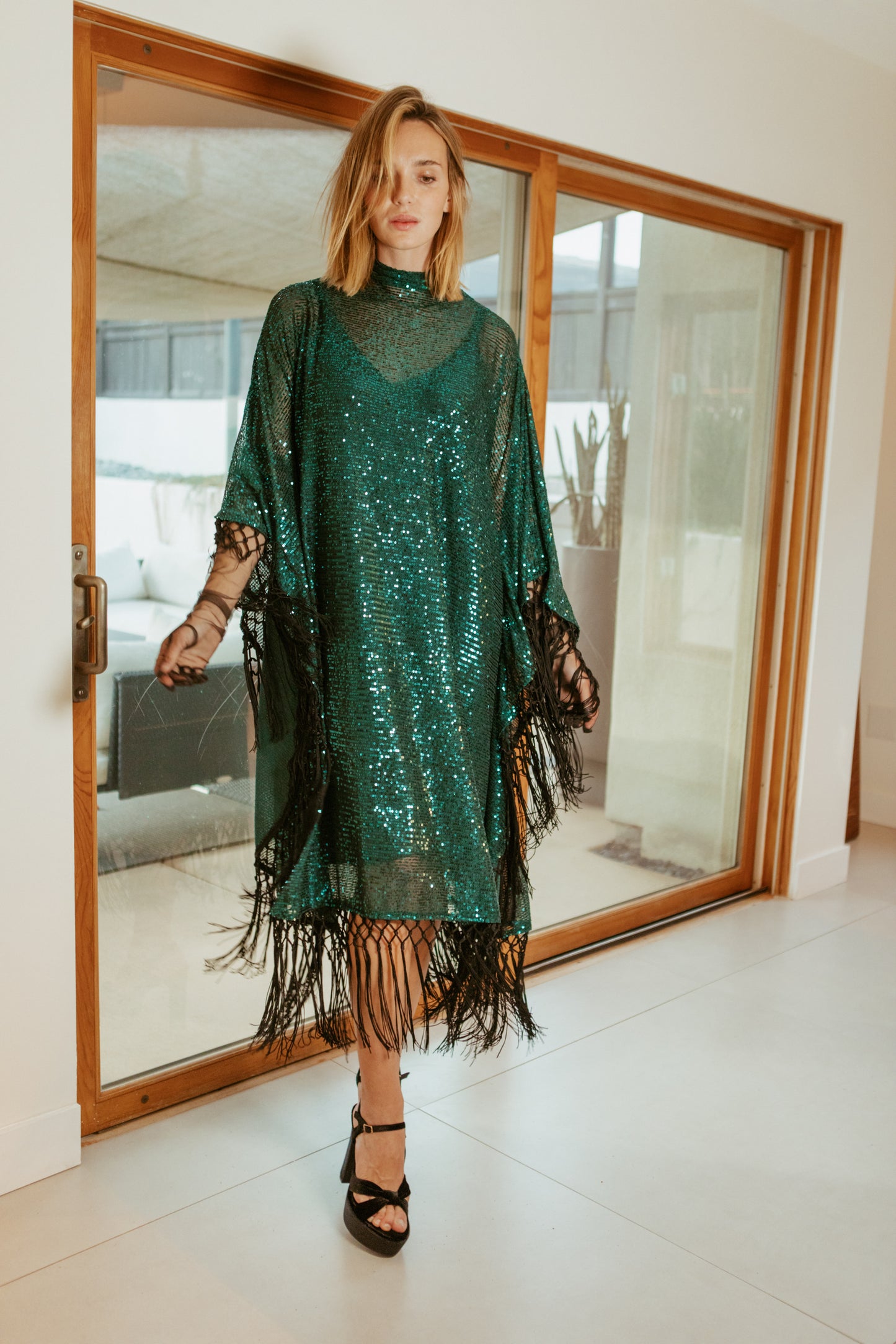 This caftan is a dazzling, full-length garment featuring a shimmering jade green sequin overlay on a mesh fabric. The sequins catch the light, creating a sparkling effect throughout. It has a mock neck collar with a straight, loose fit that drapes elegantly over the body. The edges of the caftan, including the hem and the sides, are embellished with long, silk black fringe that add movement and a playful flair.