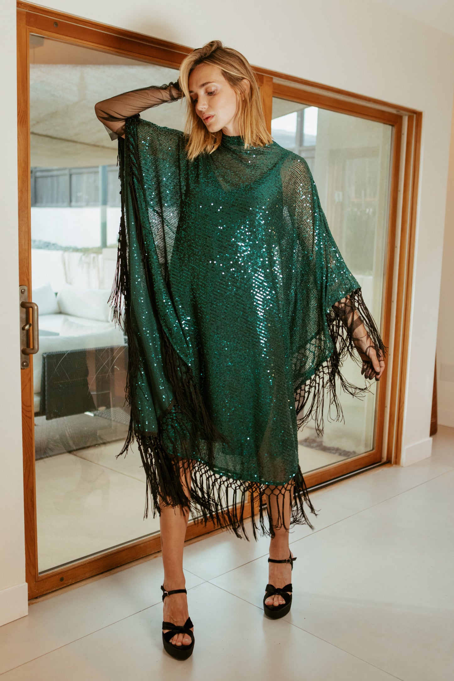 This caftan is a dazzling, full-length garment featuring a shimmering jade green sequin overlay on a mesh fabric. The sequins catch the light, creating a sparkling effect throughout. It has a mock neck collar with a straight, loose fit that drapes elegantly over the body. The edges of the caftan, including the hem and the sides, are embellished with long, silk black fringe that add movement and a playful flair.