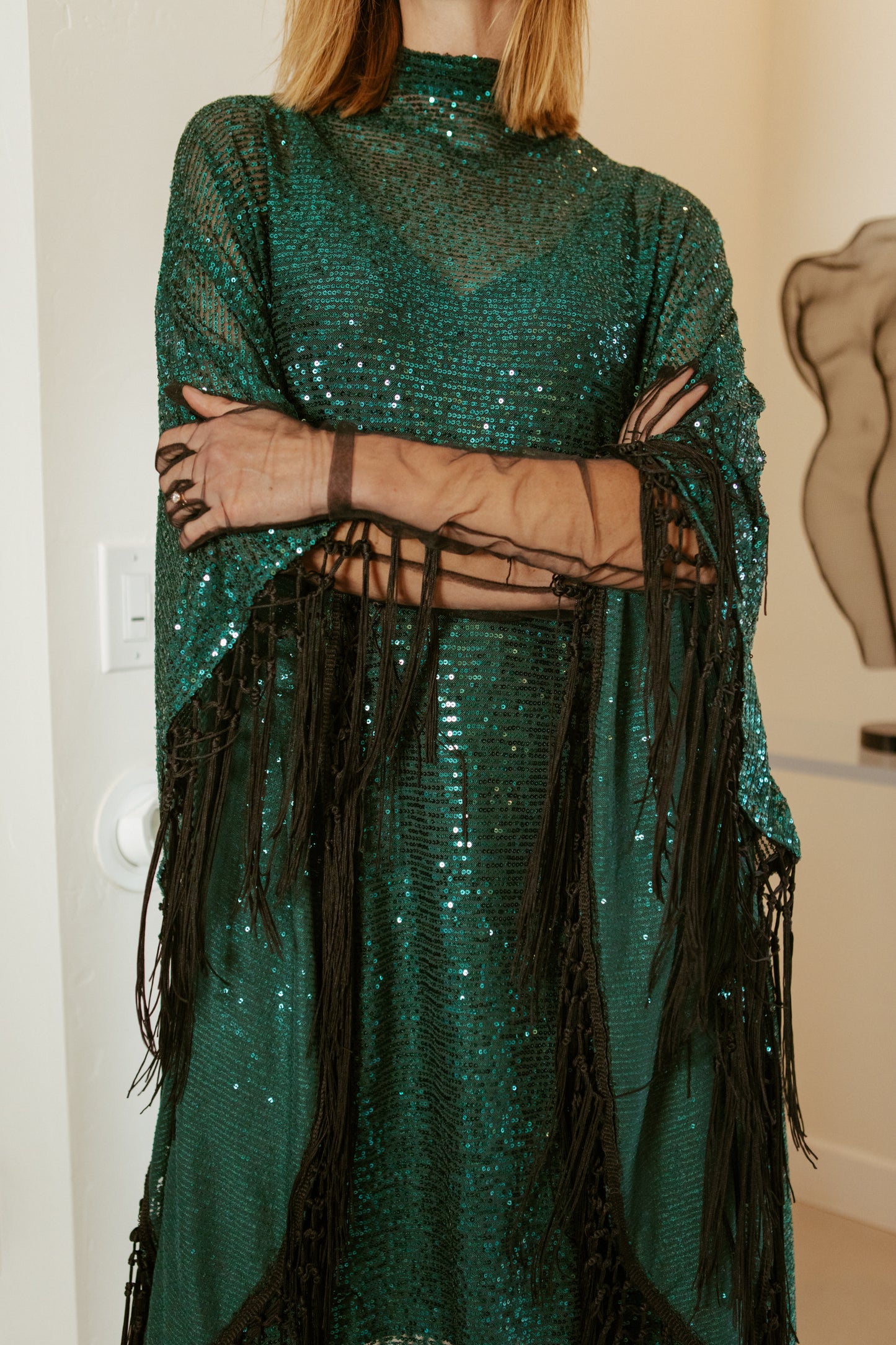 This caftan is a dazzling, full-length garment featuring a shimmering jade green sequin overlay on a mesh fabric. The sequins catch the light, creating a sparkling effect throughout. It has a mock neck collar with a straight, loose fit that drapes elegantly over the body. The edges of the caftan, including the hem and the sides, are embellished with long, silk black fringe that add movement and a playful flair.