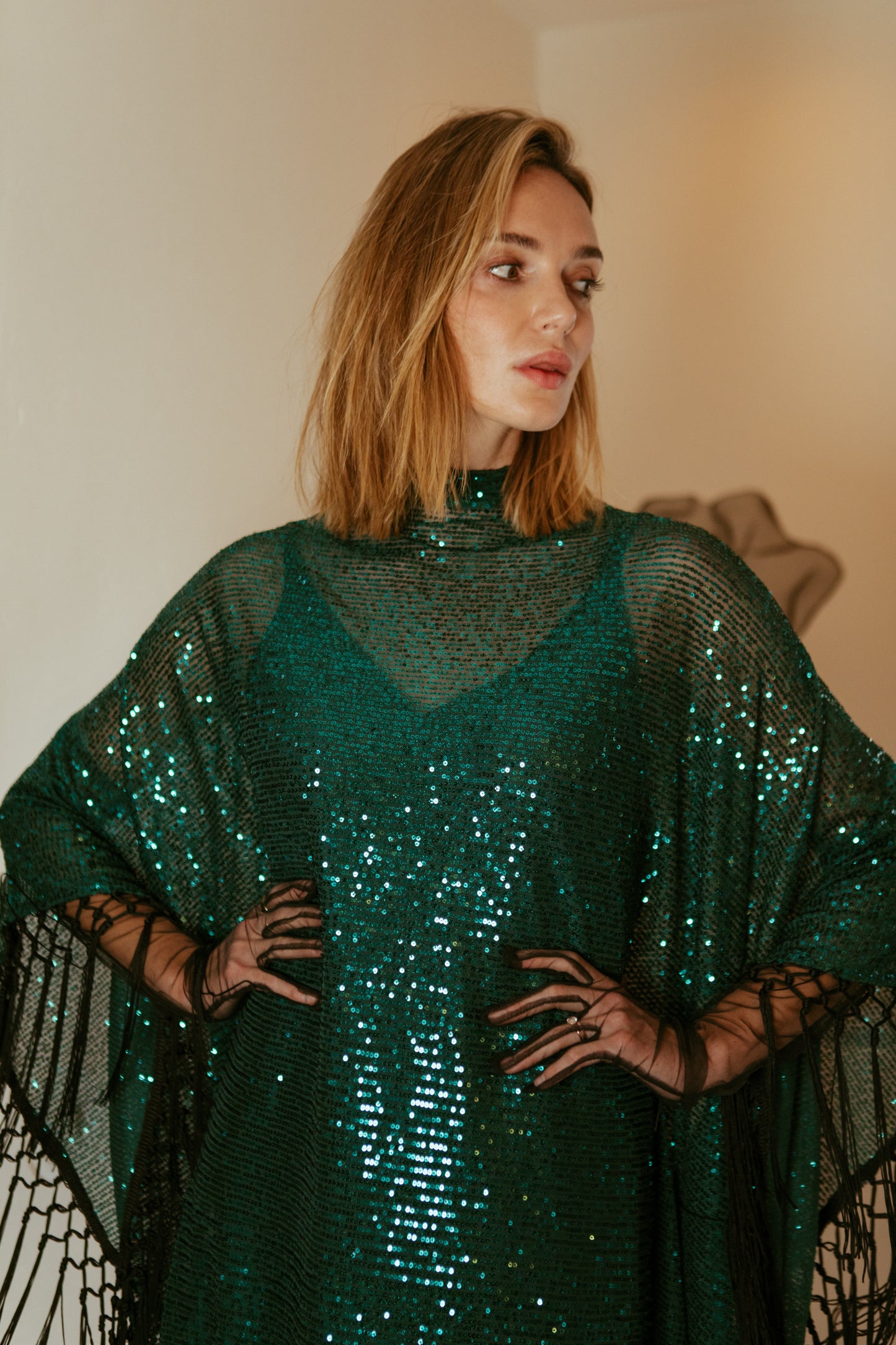 This caftan is a dazzling, full-length garment featuring a shimmering jade green sequin overlay on a mesh fabric. The sequins catch the light, creating a sparkling effect throughout. It has a mock neck collar with a straight, loose fit that drapes elegantly over the body. The edges of the caftan, including the hem and the sides, are embellished with long, silk black fringe that add movement and a playful flair.