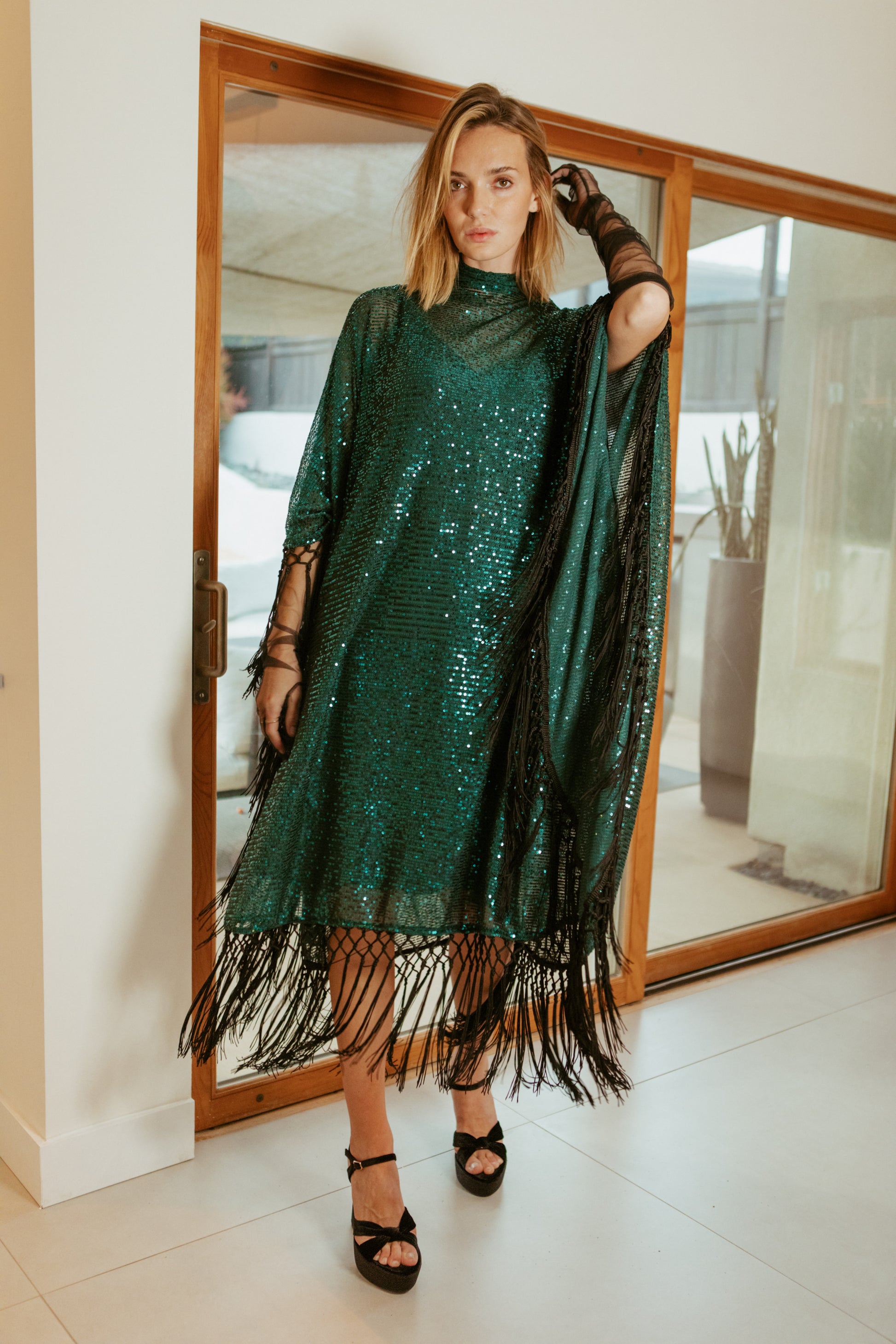 This caftan is a dazzling, full-length garment featuring a shimmering jade green sequin overlay on a mesh fabric. The sequins catch the light, creating a sparkling effect throughout. It has a mock neck collar with a straight, loose fit that drapes elegantly over the body. The edges of the caftan, including the hem and the sides, are embellished with long, silk black fringe that add movement and a playful flair.