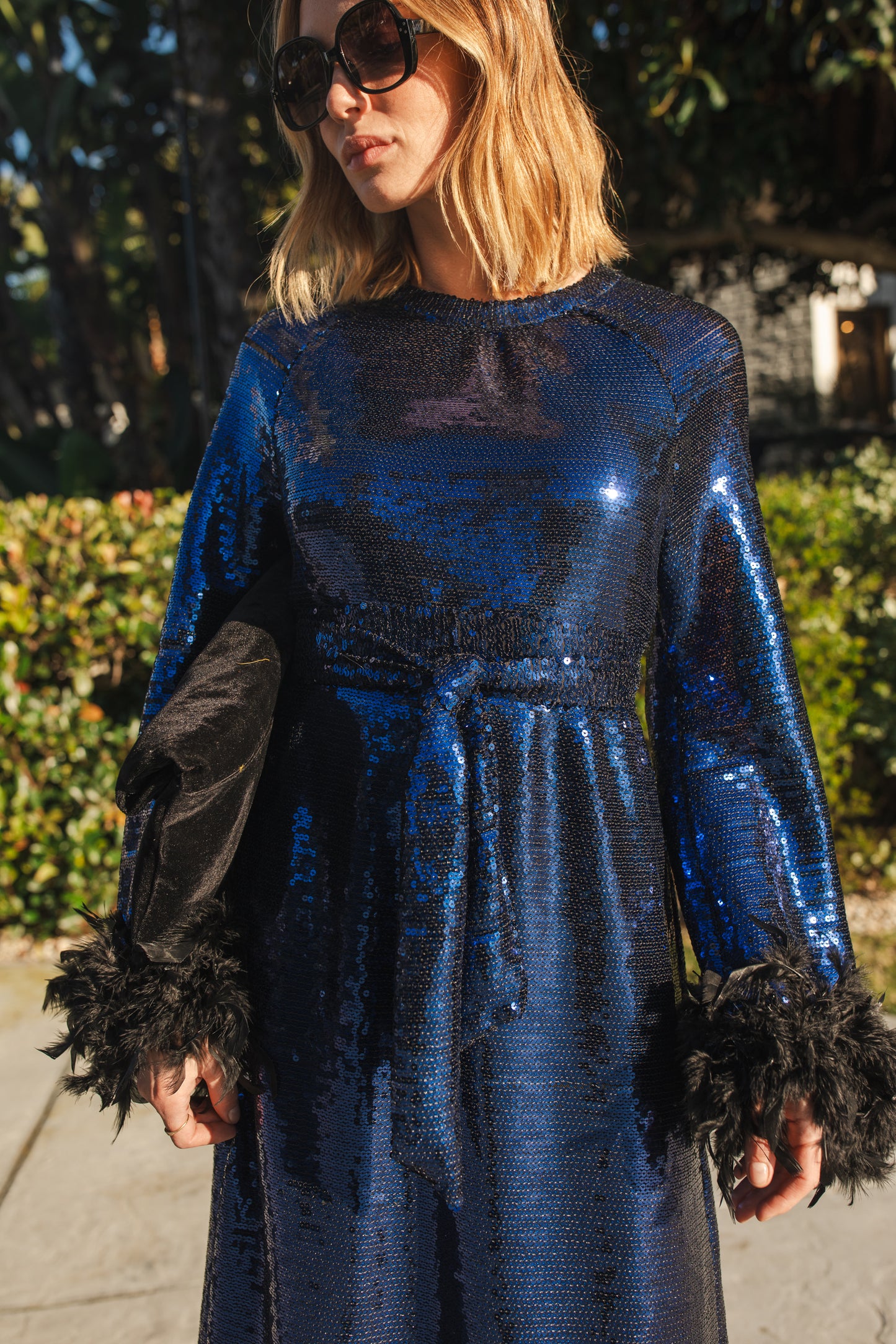 Model wears a knee length dress covered in midnight blue sequins, the dress has a crew neck and full long sleeves with a feather trim.