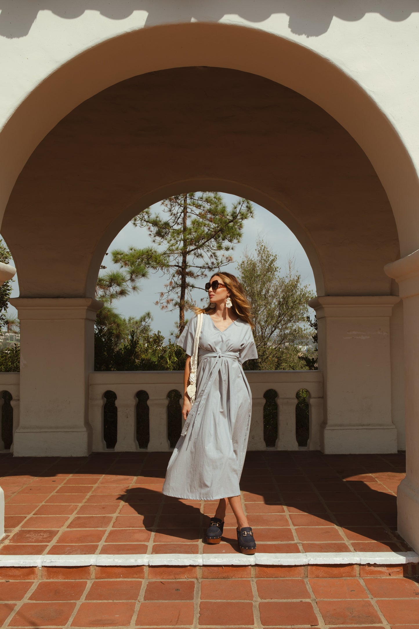 White and blue vertical pinstripe a-line midi-maxi dress featuring v-neck, short sleeves, and built-in sash ties for a cinched, wrap waist look. This dress falls at approximately lower calf or ankle. Nautical bohemian in style.