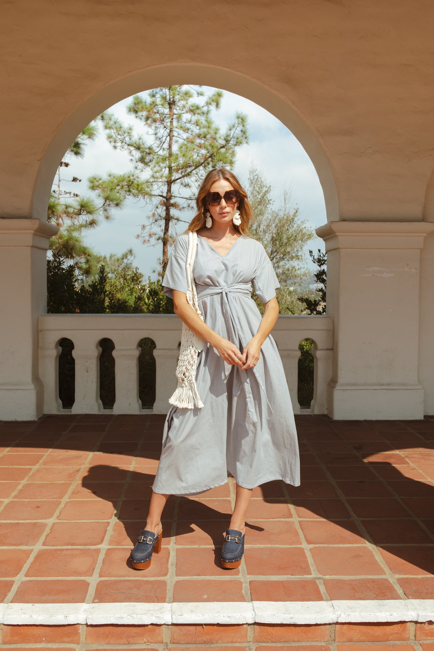 White and blue vertical pinstripe a-line midi-maxi dress featuring v-neck, short sleeves, and built-in sash ties for a cinched, wrap waist look. This dress falls at approximately lower calf or ankle. Nautical bohemian in style.