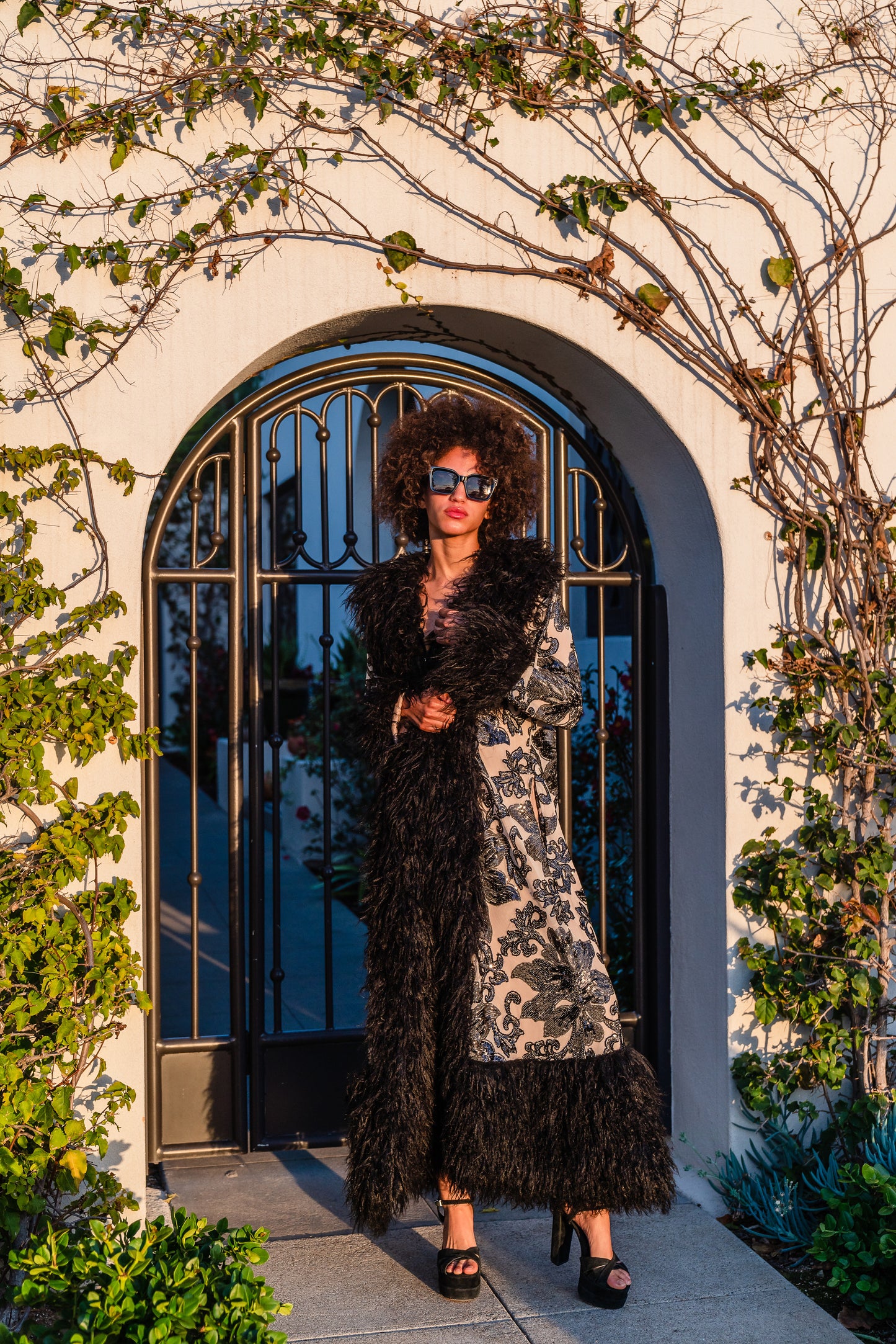 A floor length coat made of nude mesh with black sequins in a floral patter. The coat is lined around the collar, down the opening and around the hem in fluffy black faux alpaca fur, the coat has pockets and a nude colored lining of bamboo jersey.