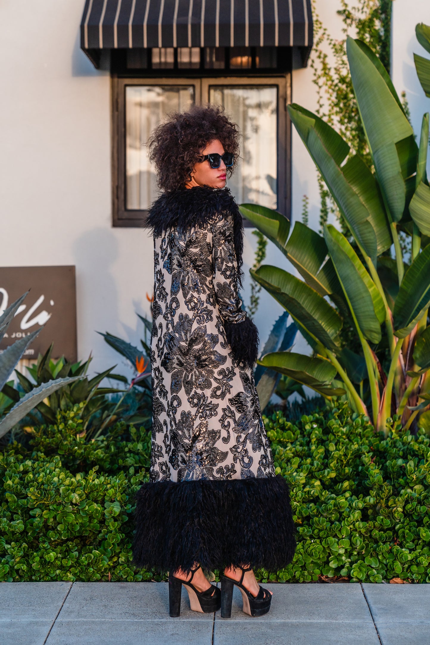 A floor length coat made of nude mesh with black sequins in a floral patter. The coat is lined around the collar, down the opening and around the hem in fluffy black faux alpaca fur, the coat has pockets and a nude colored lining of bamboo jersey.