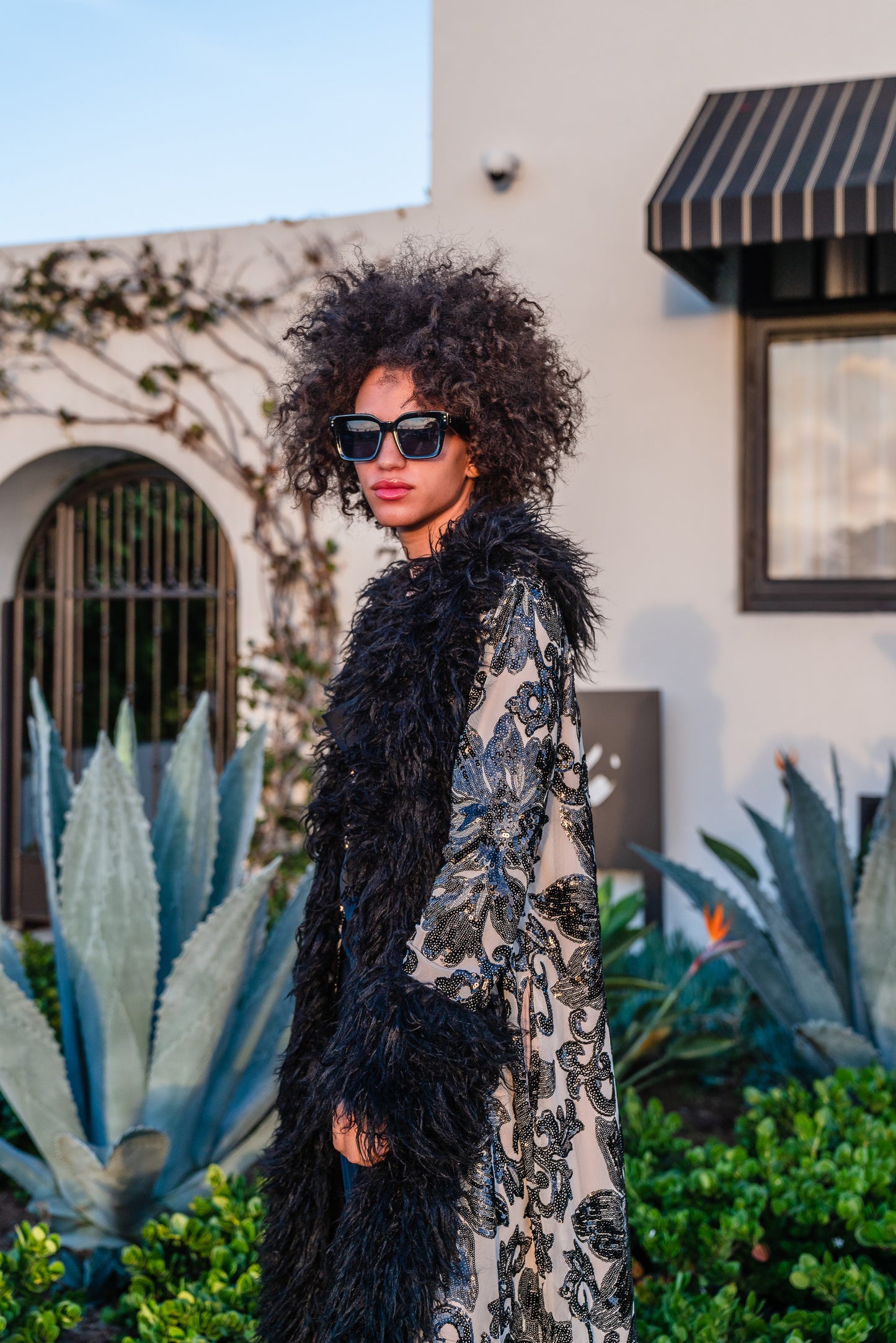 A floor length coat made of nude mesh with black sequins in a floral patter. The coat is lined around the collar, down the opening and around the hem in fluffy black faux alpaca fur, the coat has pockets and a nude colored lining of bamboo jersey.