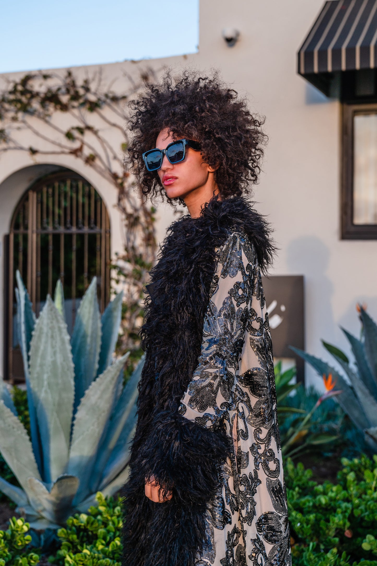 A floor length coat made of nude mesh with black sequins in a floral patter. The coat is lined around the collar, down the opening and around the hem in fluffy black faux alpaca fur, the coat has pockets and a nude colored lining of bamboo jersey.