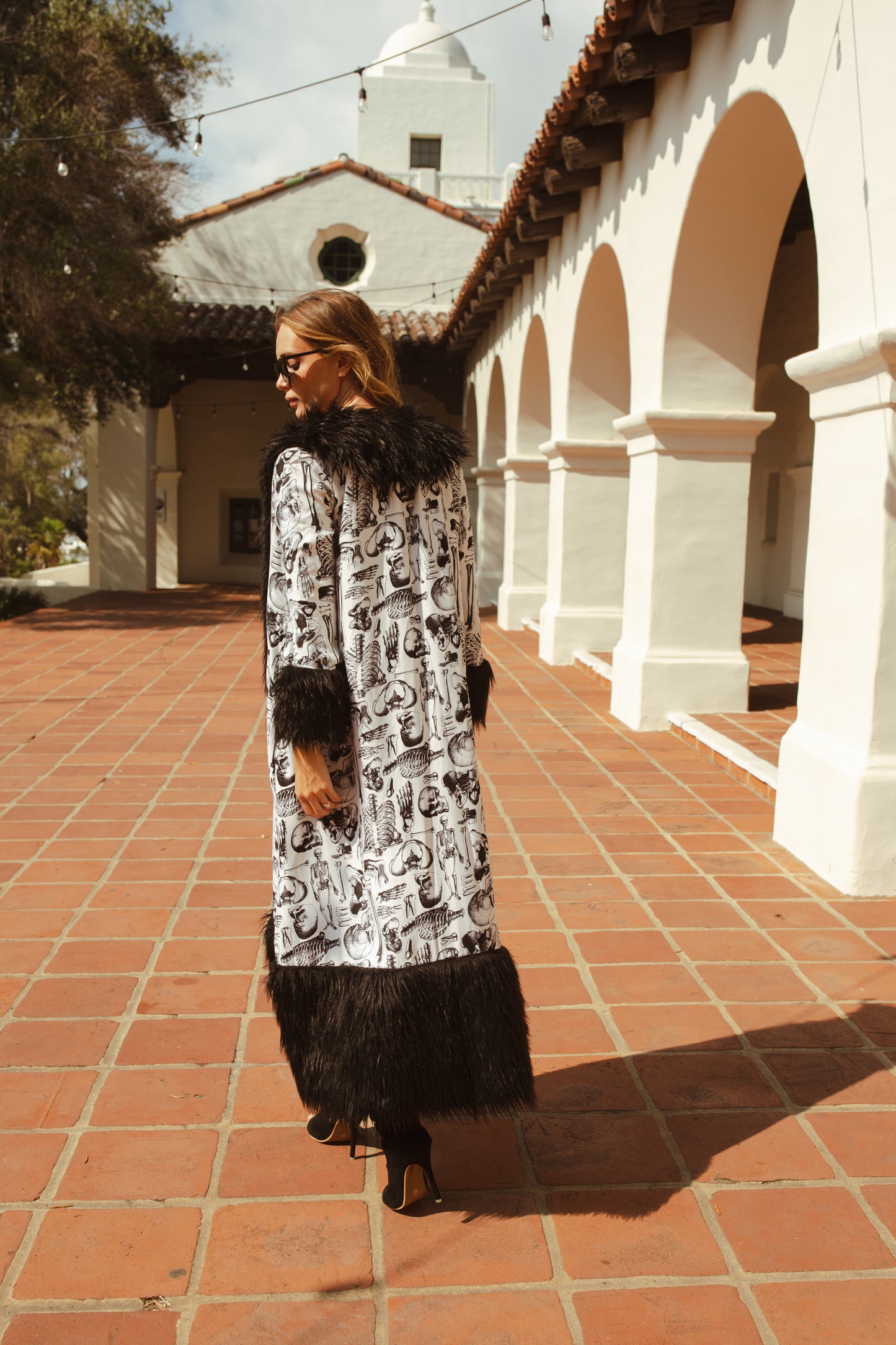 Long, penny lane style trench coat fashioned from an ivory white fabric overlaid with anatomical skeleton sketches. Embellished with long, black faux fur along collar, lapel, cuffs, and hem. High contrast black and white, spooky gothic aesthetic mixed with retro 70s style.