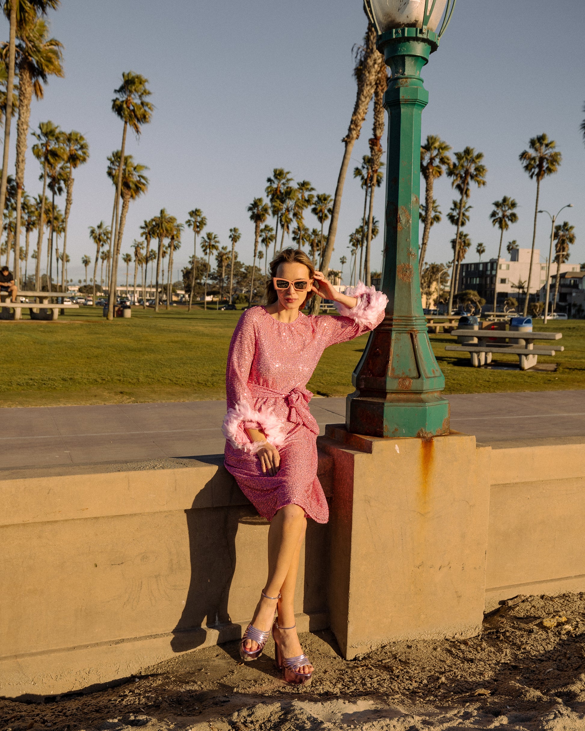 Jennafer Grace light pink flamingo sequins dress. Featuring a boat neck, generous bishop sleeves with light pink feather detail at sleeve hem, flowing midi length maxi skirt, and a slim matching sash for a cinched waist.