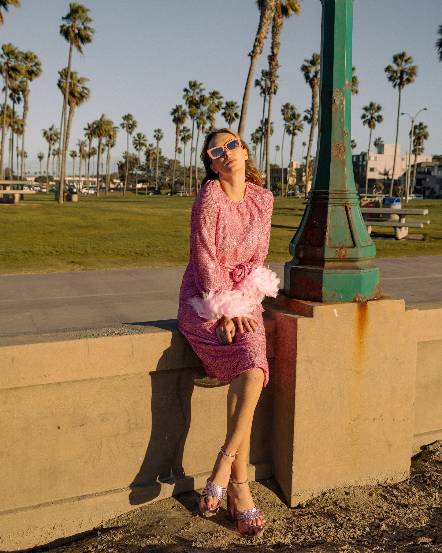 Jennafer Grace light pink flamingo sequins dress. Featuring a boat neck, generous bishop sleeves with light pink feather detail at sleeve hem, flowing midi length maxi skirt, and a slim matching sash for a cinched waist.