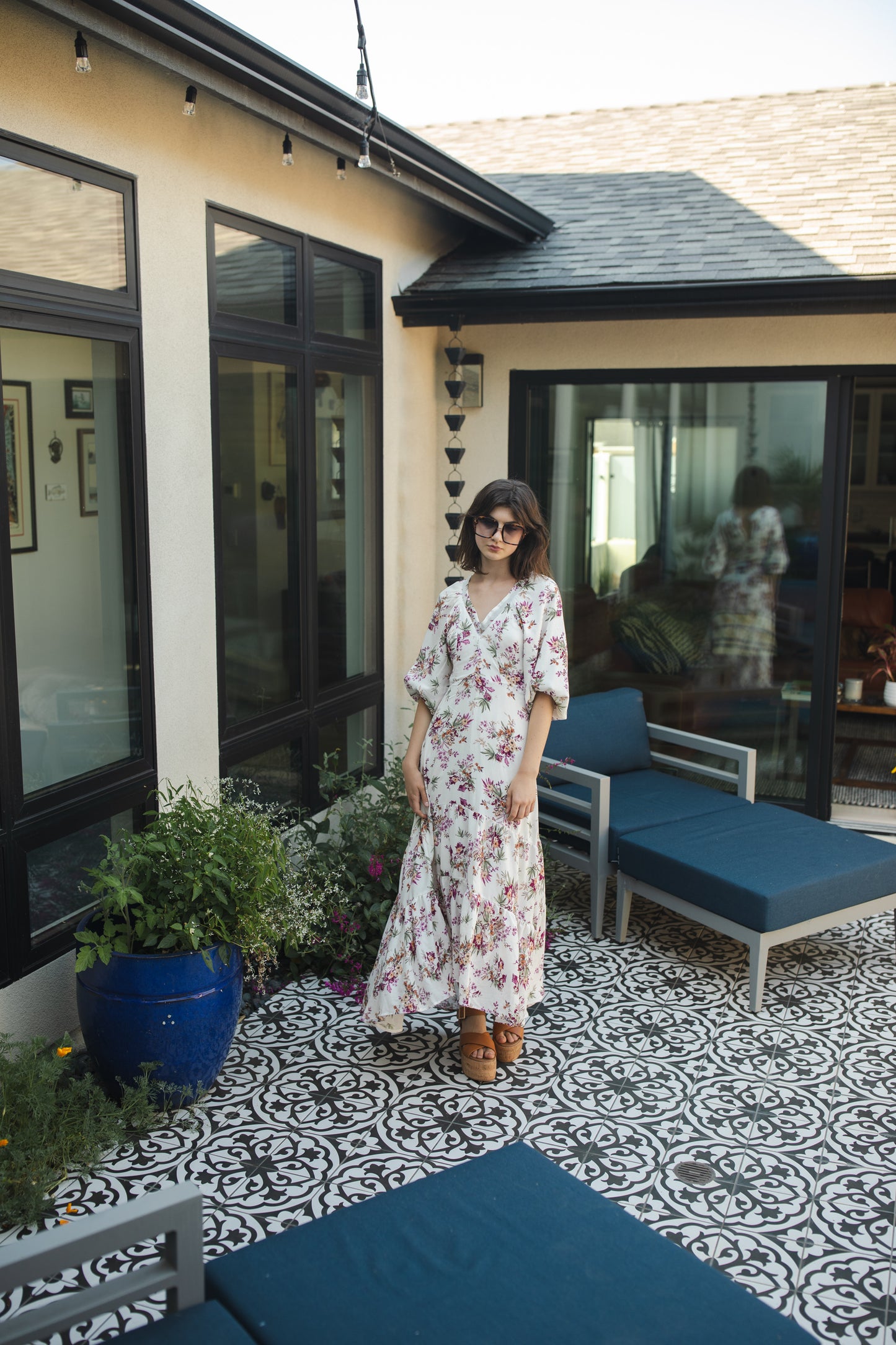 The Vana Vixen maxi dress by Jennafer Grace with a white base and summer floral print of deep pinks, yellows, and greens. Made from a linen like cotton rayon frabric, featuring a v-neck, detailed bishop sleeves, tiered ruffle skirt with ankle length hem, and a keyhole slit at back.