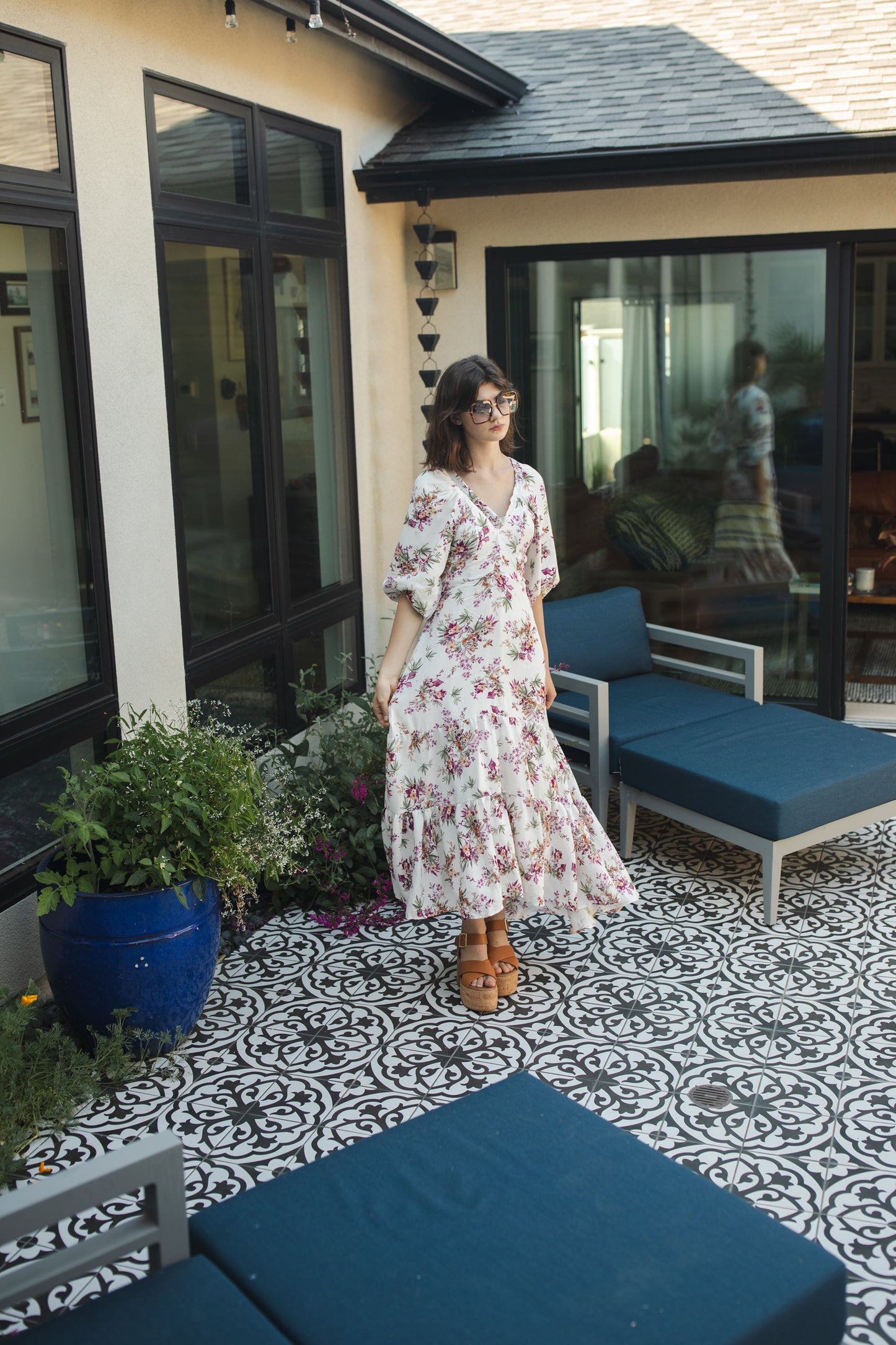 The Vana Vixen maxi dress by Jennafer Grace with a white base and summer floral print of deep pinks, yellows, and greens. Made from a linen like cotton rayon frabric, featuring a v-neck, detailed bishop sleeves, tiered ruffle skirt with ankle length hem, and a keyhole slit at back.