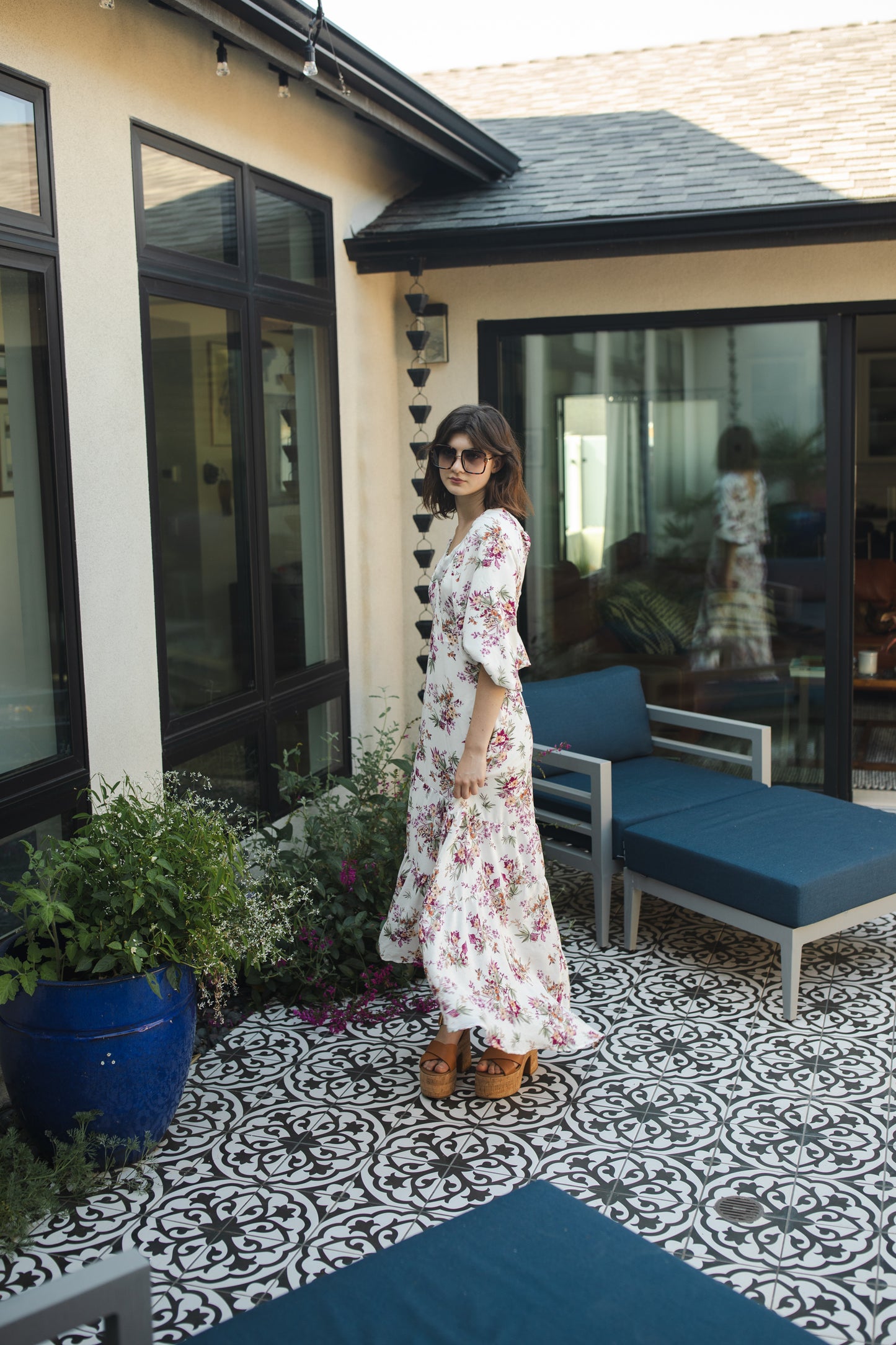 The Vana Vixen maxi dress by Jennafer Grace with a white base and summer floral print of deep pinks, yellows, and greens. Made from a linen like cotton rayon frabric, featuring a v-neck, detailed bishop sleeves, tiered ruffle skirt with ankle length hem, and a keyhole slit at back.
