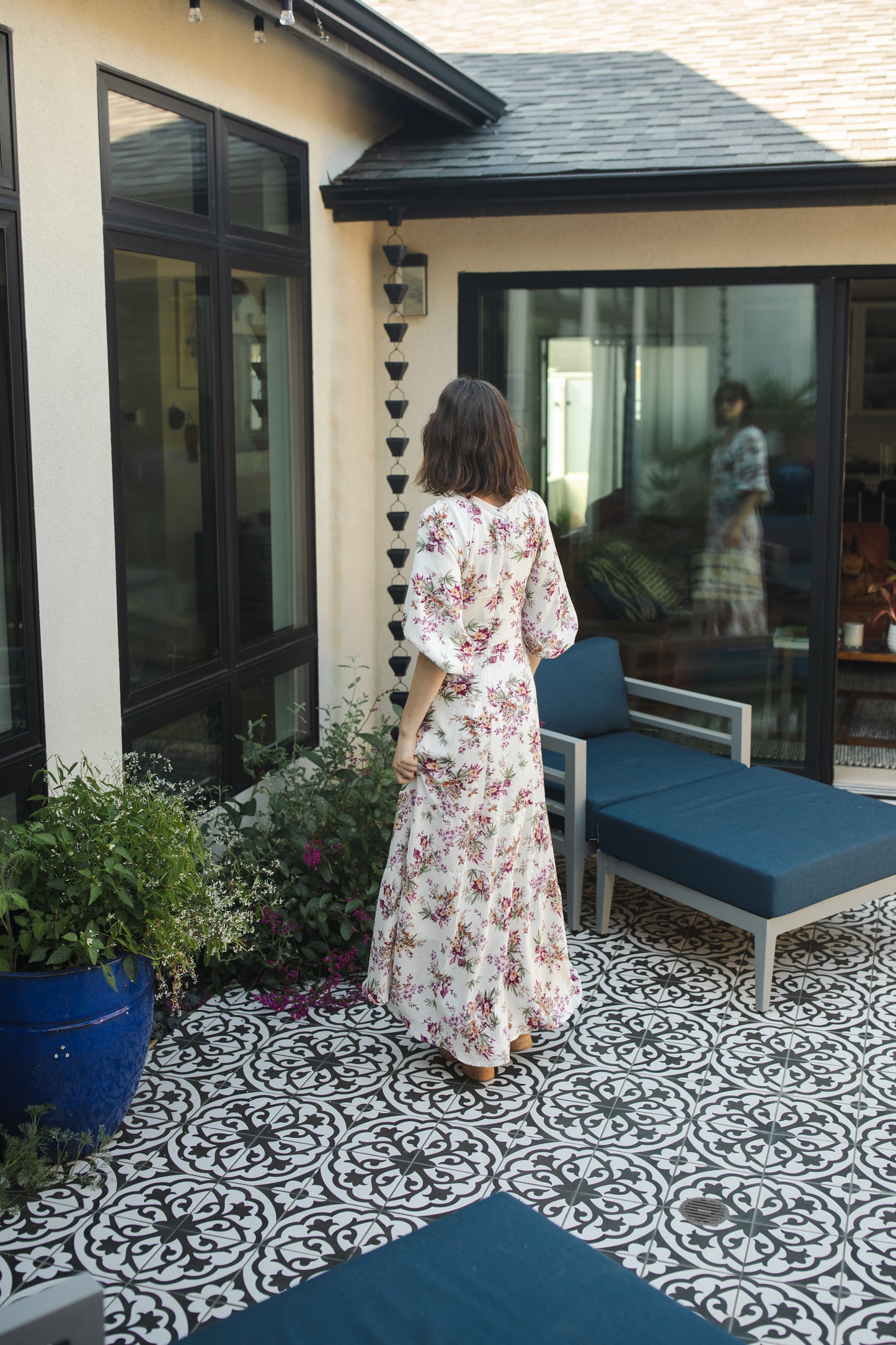 The Vana Vixen maxi dress by Jennafer Grace with a white base and summer floral print of deep pinks, yellows, and greens. Made from a linen like cotton rayon frabric, featuring a v-neck, detailed bishop sleeves, tiered ruffle skirt with ankle length hem, and a keyhole slit at back.