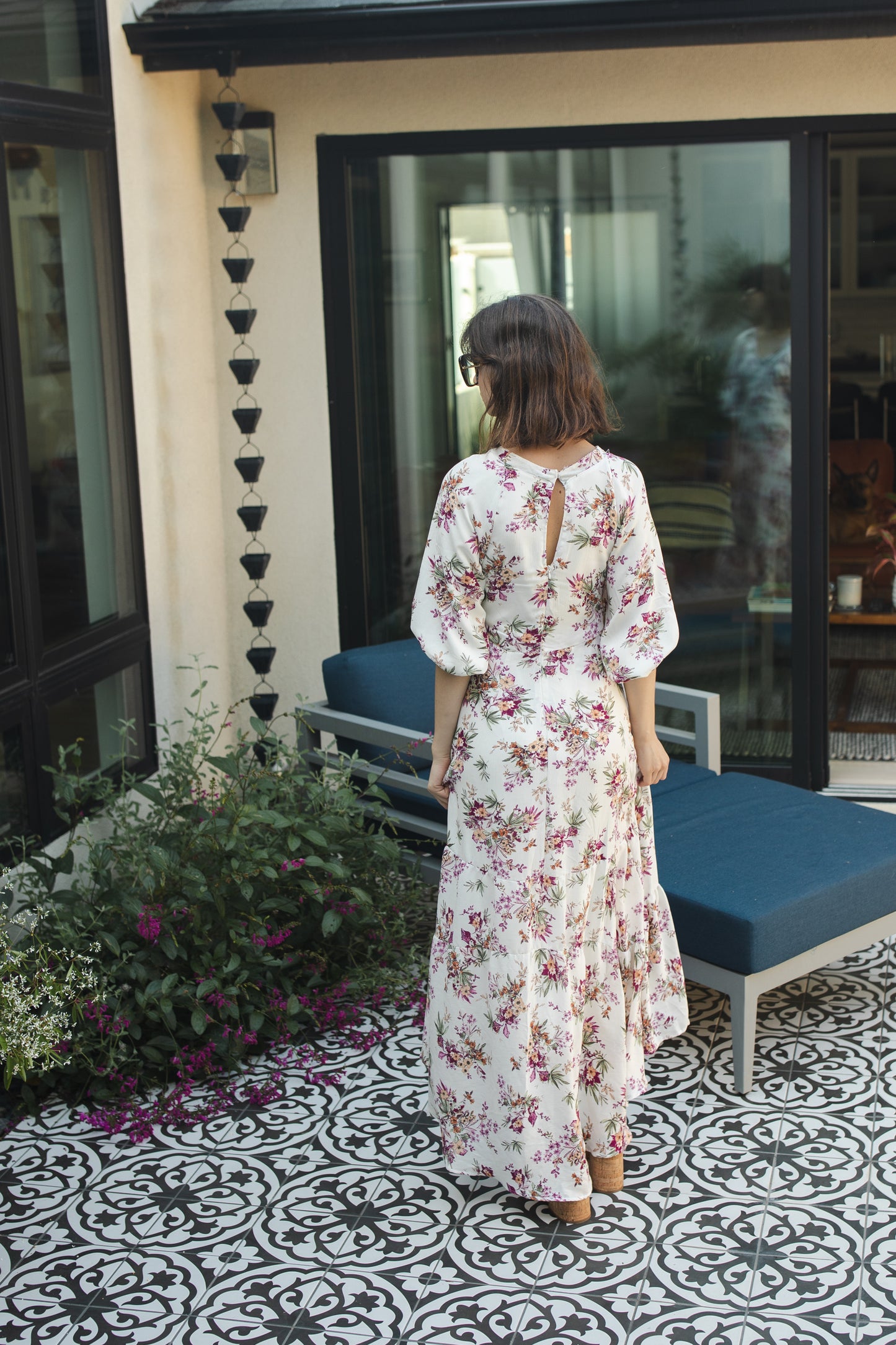 The Vana Vixen maxi dress by Jennafer Grace with a white base and summer floral print of deep pinks, yellows, and greens. Made from a linen like cotton rayon frabric, featuring a v-neck, detailed bishop sleeves, tiered ruffle skirt with ankle length hem, and a keyhole slit at back.