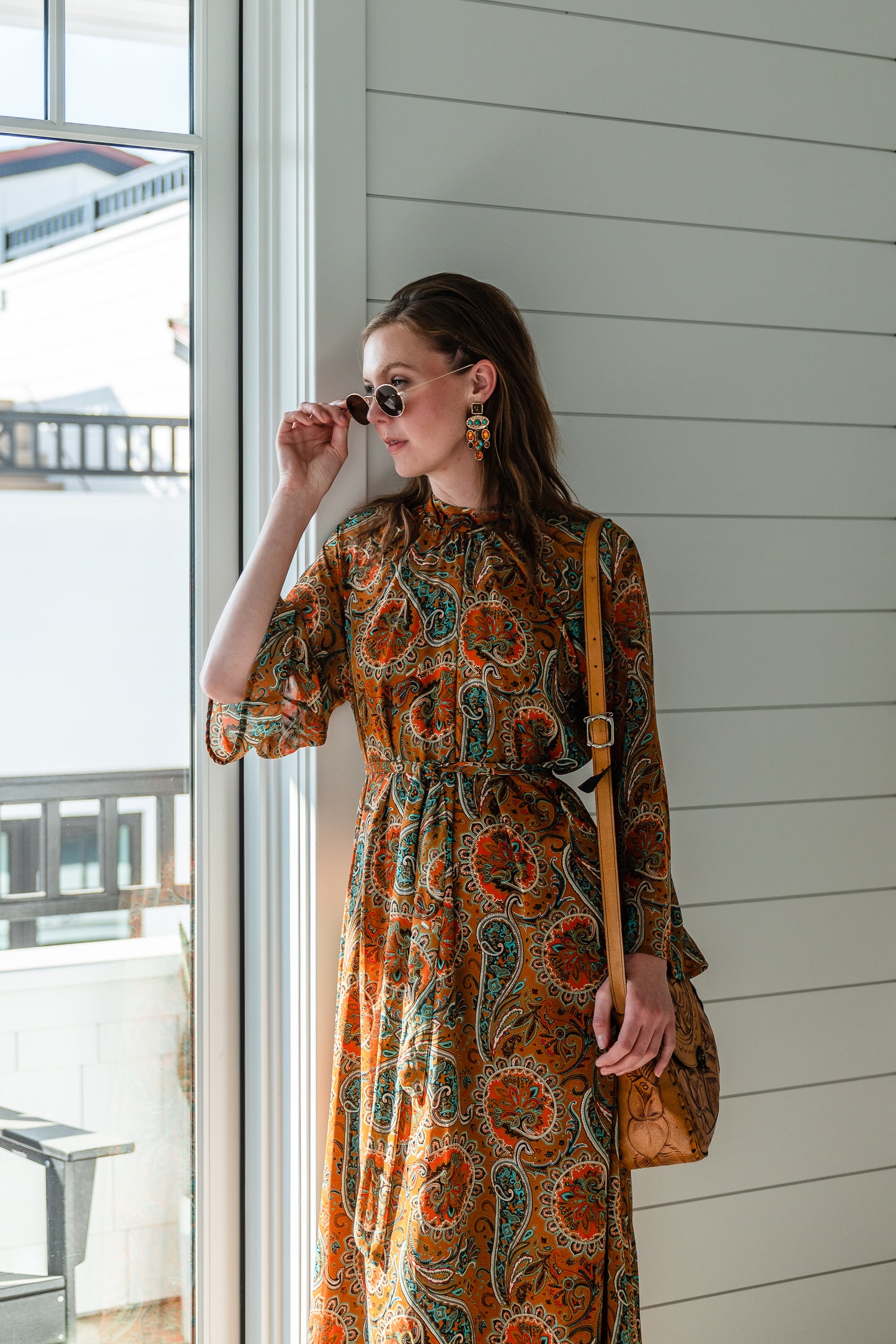 Model wears a soft chiffon midi length dress in caramel brown with turquoise, green and orange paisley print. The sleeves are bell shaped and the dress has a wrap around belt
