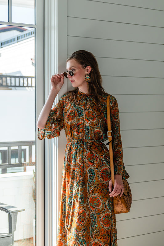 Model wears a soft chiffon midi length dress in caramel brown with turquoise, green and orange paisley print. The sleeves are bell shaped and the dress has a wrap around belt