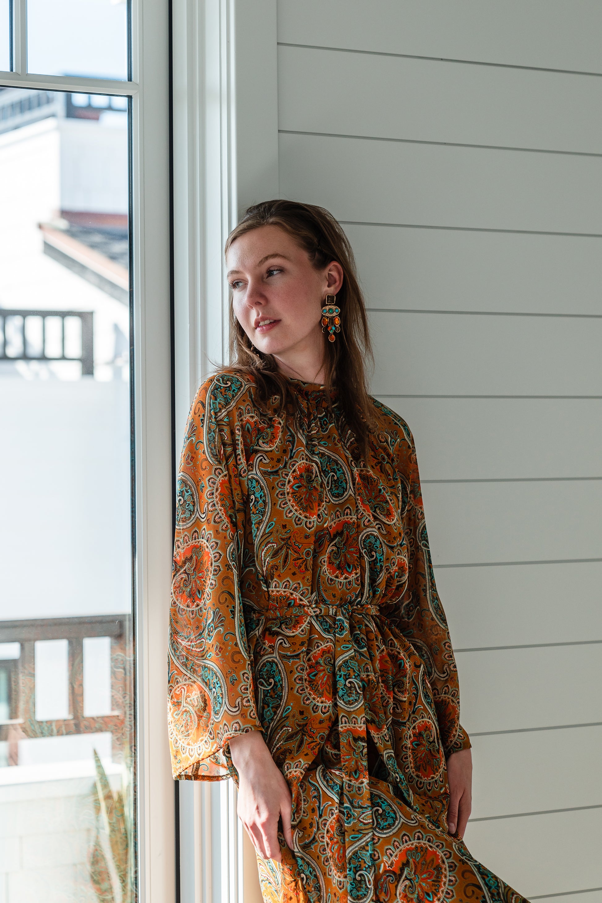 Model wears a soft chiffon midi length dress in caramel brown with turquoise, green and orange paisley print. The sleeves are bell shaped and the dress has a wrap around belt