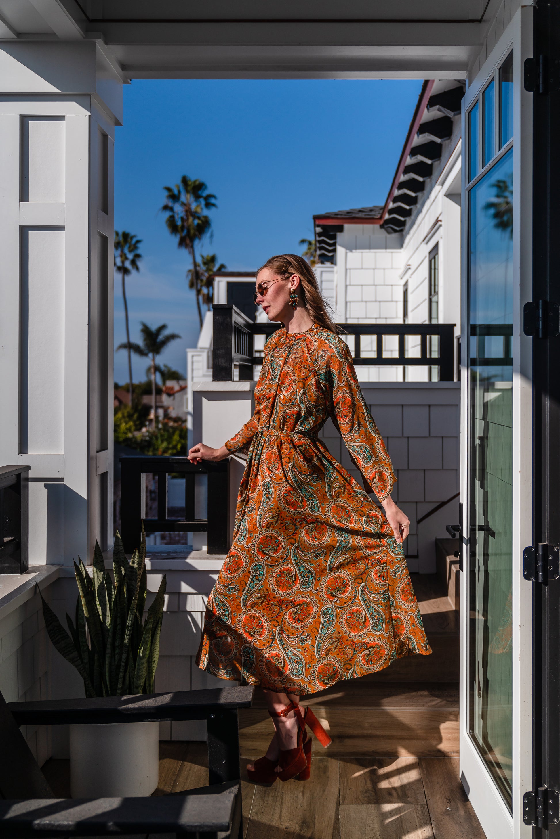 Model wears a soft chiffon midi length dress in caramel brown with turquoise, green and orange paisley print. The sleeves are bell shaped and the dress has a wrap around belt