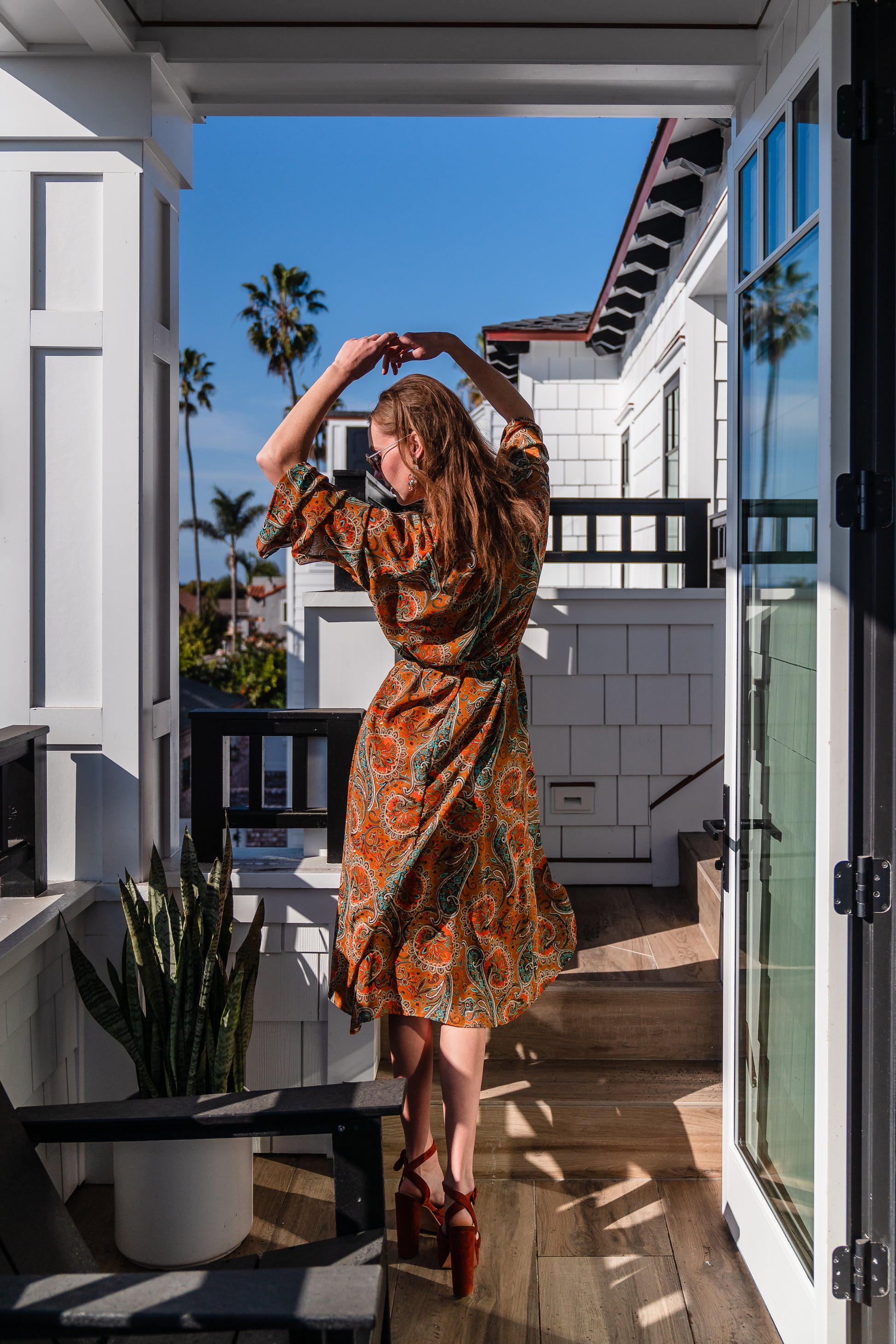 Model wears a soft chiffon midi length dress in caramel brown with turquoise, green and orange paisley print. The sleeves are bell shaped and the dress has a wrap around belt