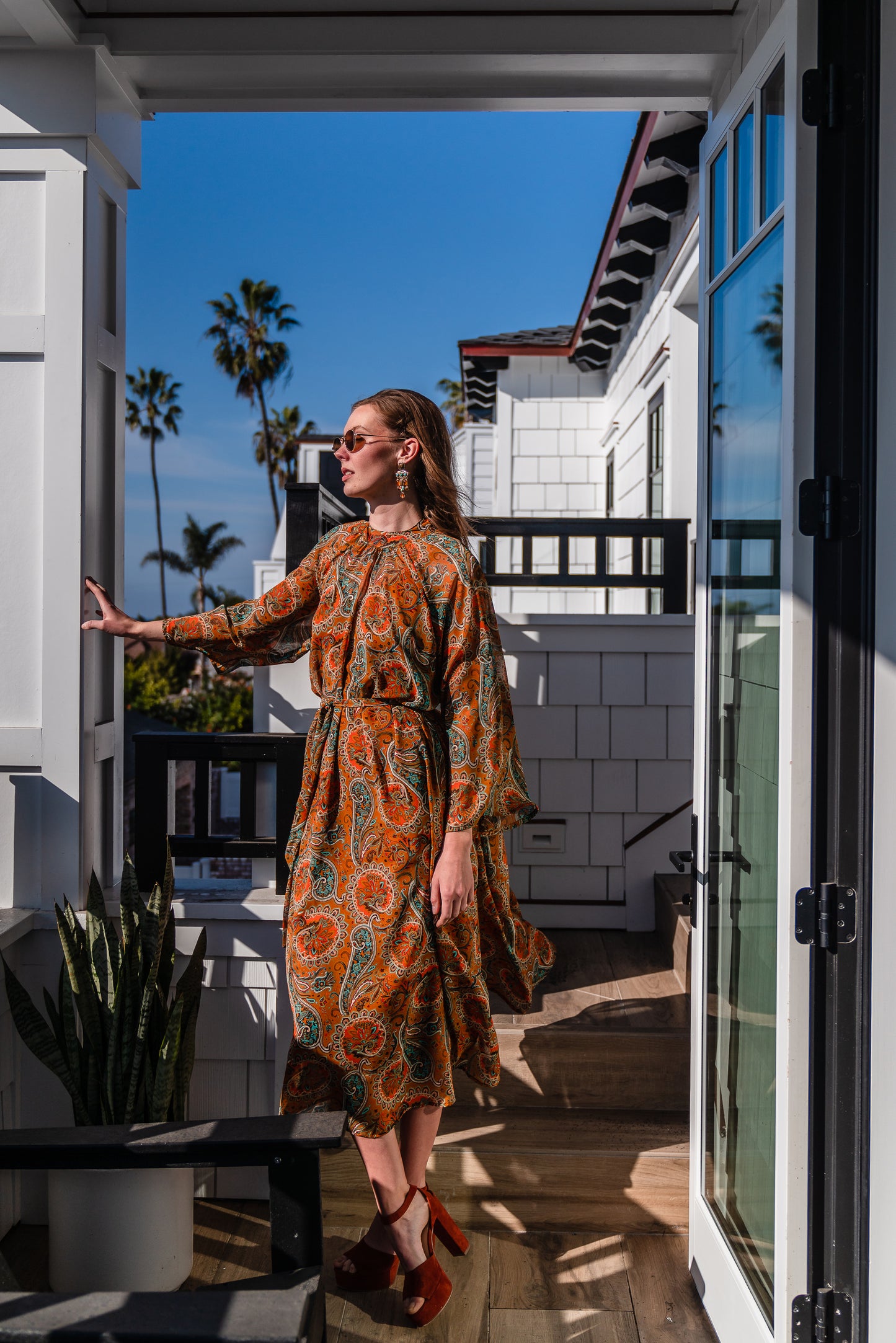 Model wears a soft chiffon midi length dress in caramel brown with turquoise, green and orange paisley print. The sleeves are bell shaped and the dress has a wrap around belt