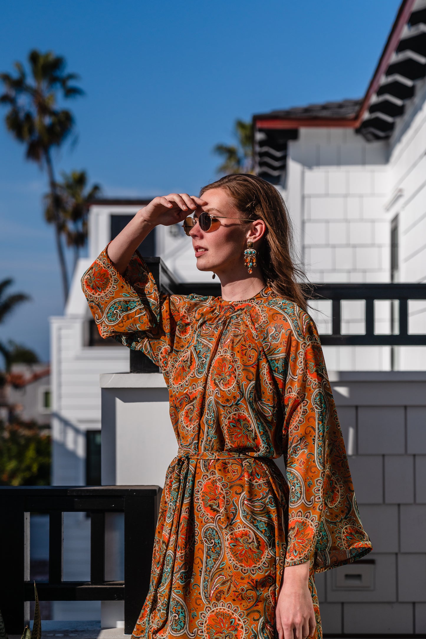 Model wears a soft chiffon midi length dress in caramel brown with turquoise, green and orange paisley print. The sleeves are bell shaped and the dress has a wrap around belt