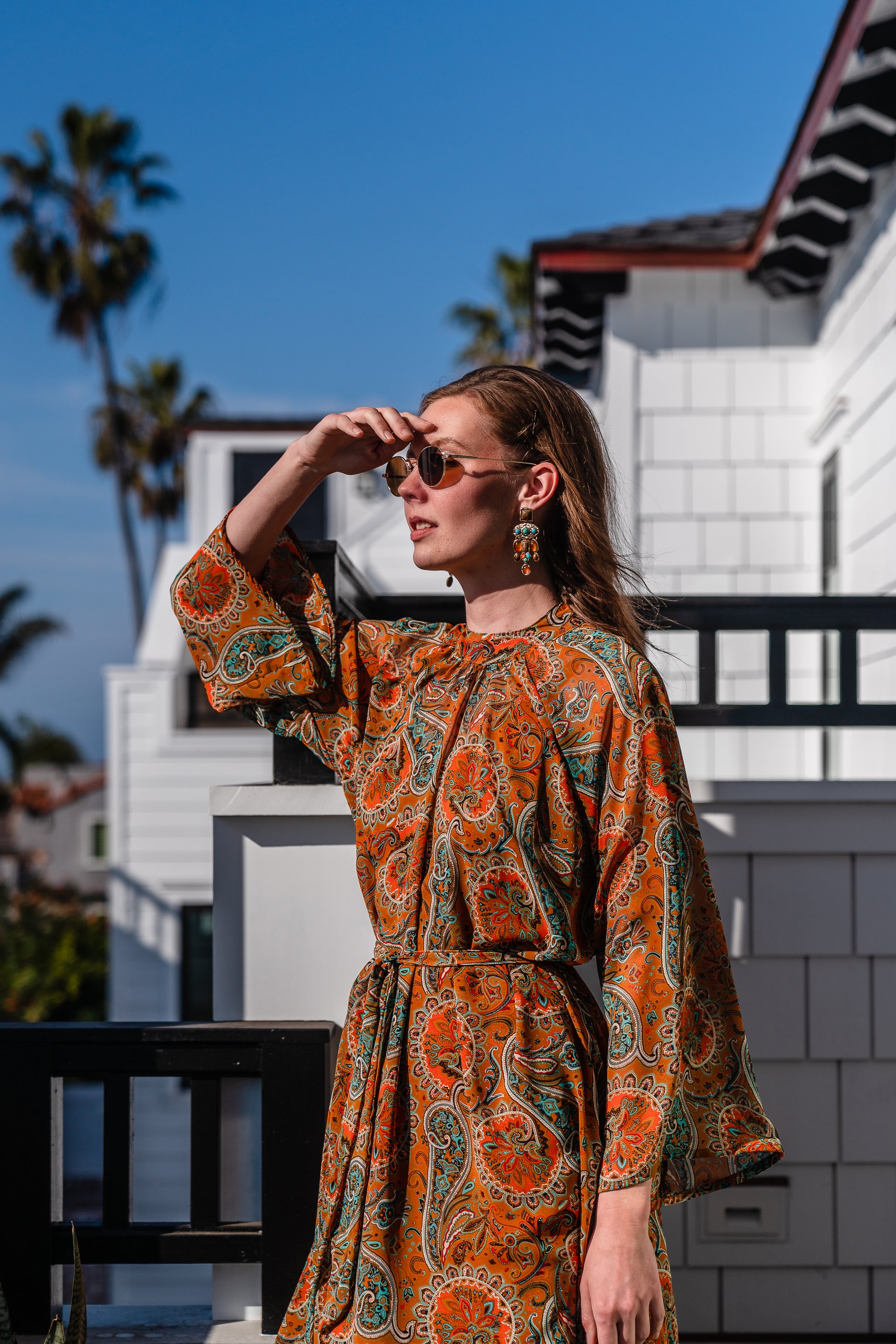 Model wears a soft chiffon midi length dress in caramel brown with turquoise, green and orange paisley print. The sleeves are bell shaped and the dress has a wrap around belt