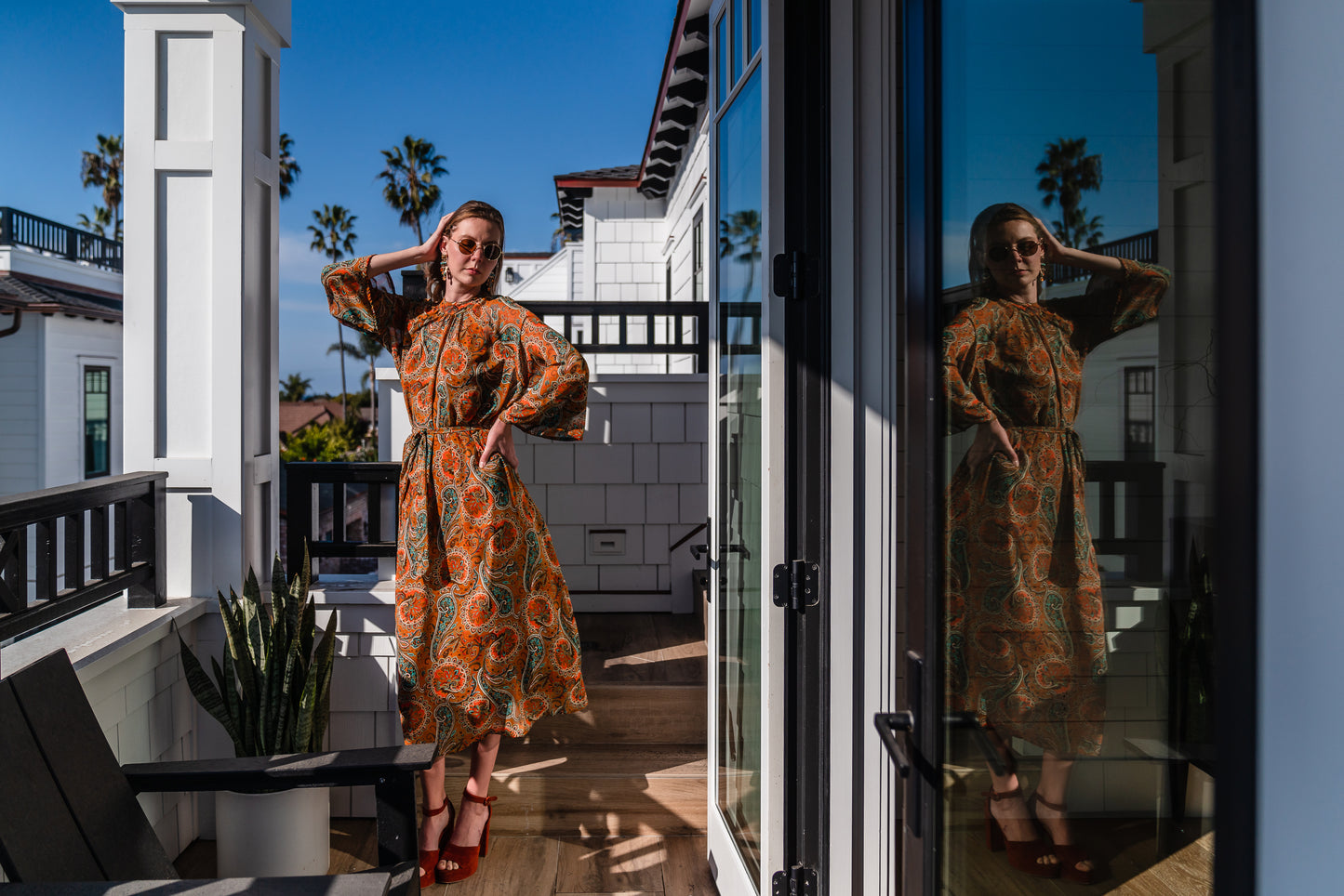 Model wears a soft chiffon midi length dress in caramel brown with turquoise, green and orange paisley print. The sleeves are bell shaped and the dress has a wrap around belt