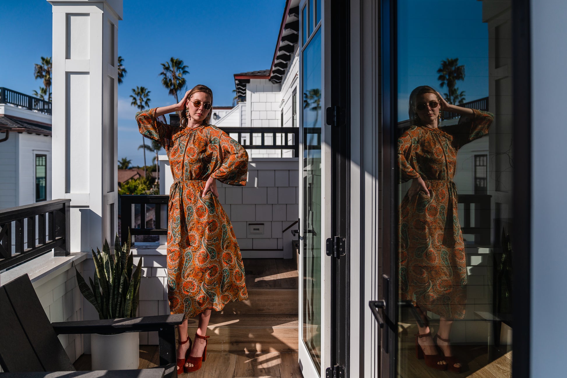 Model wears a soft chiffon midi length dress in caramel brown with turquoise, green and orange paisley print. The sleeves are bell shaped and the dress has a wrap around belt
