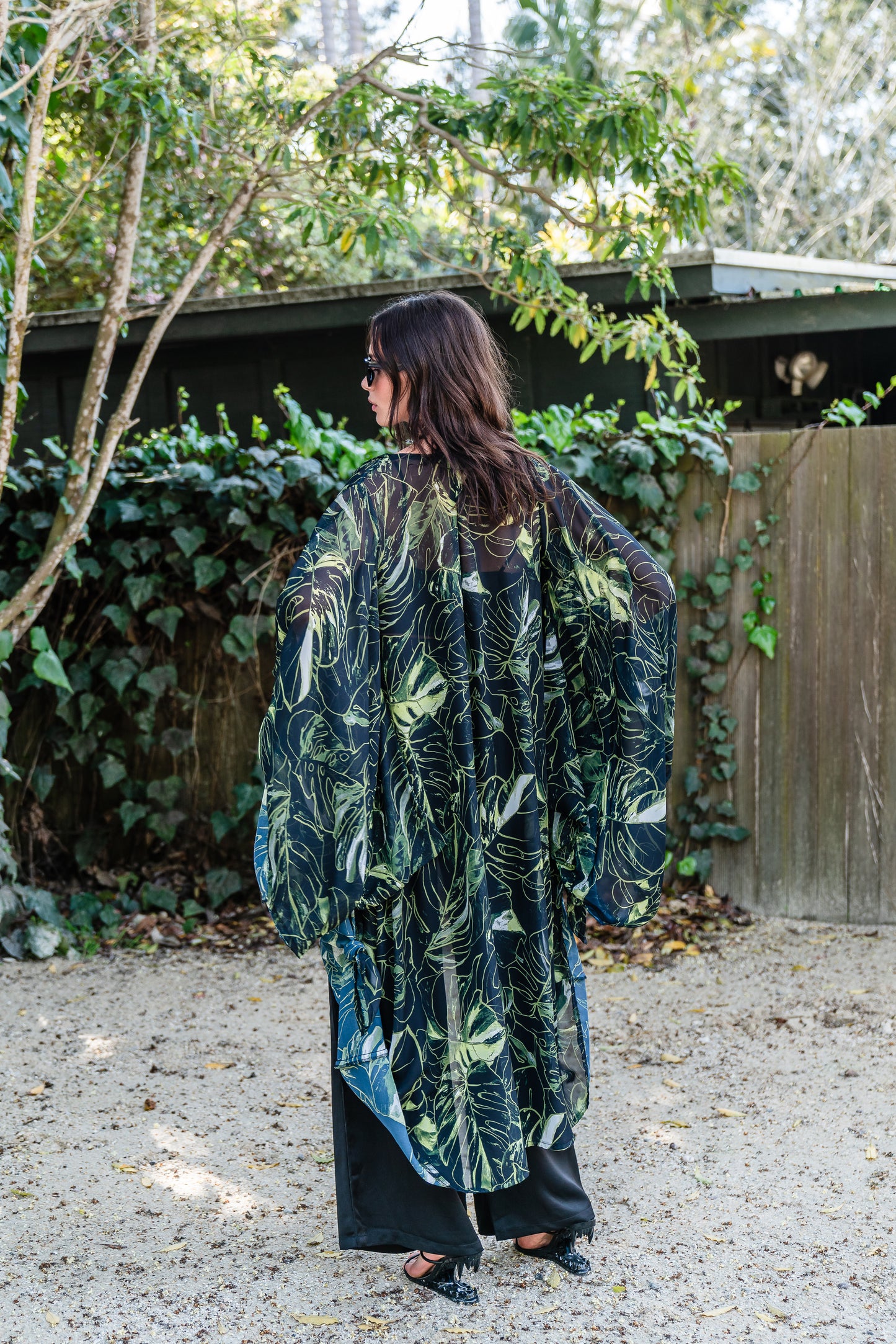 Model wears black chiffon Kimono with varying green palm leaf print. The kimono has classic wide kimono sleeves and lands at midi length. Kimono has a matching tie belt.