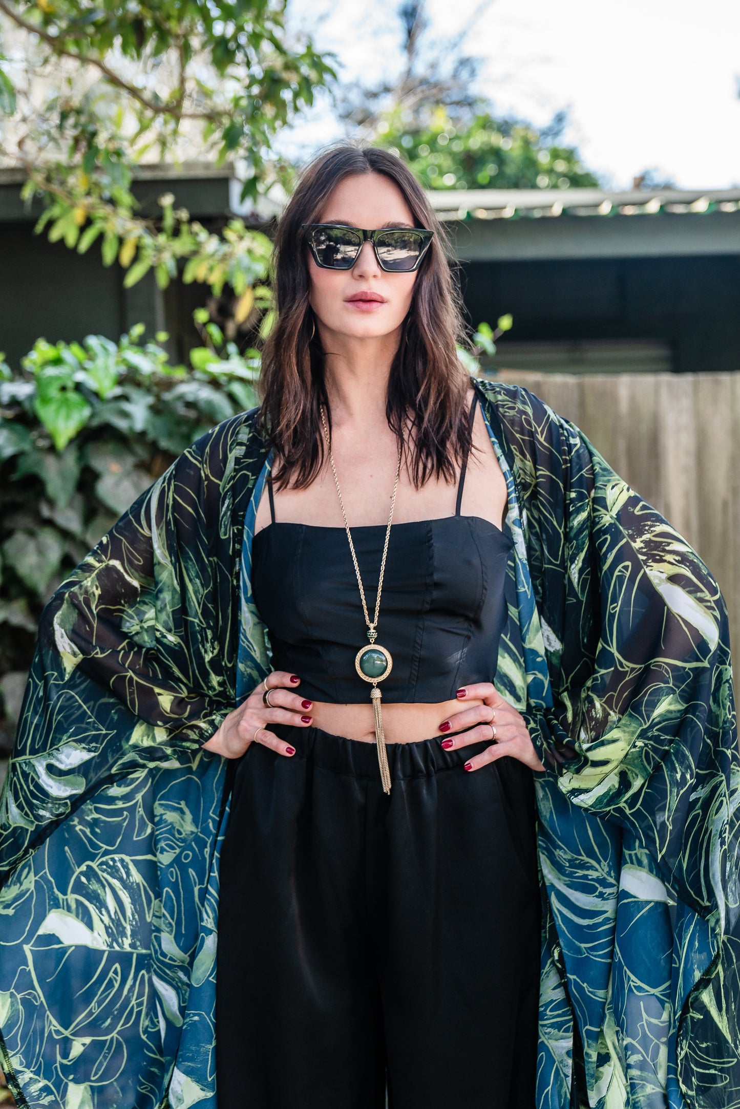 Model wears black chiffon Kimono with varying green palm leaf print. The kimono has classic wide kimono sleeves and lands at midi length. Kimono has a matching tie belt.