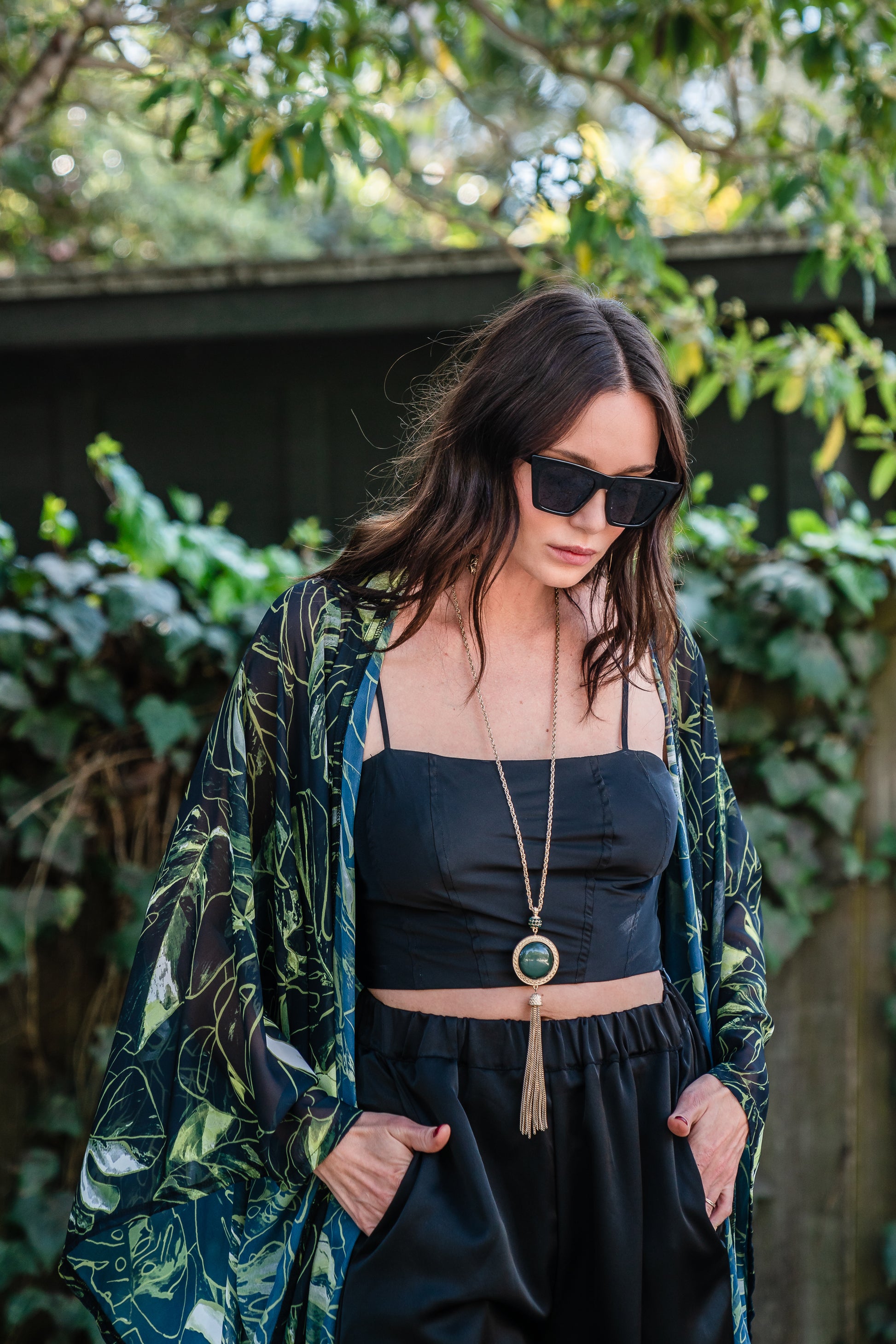 Model wears black chiffon Kimono with varying green palm leaf print. The kimono has classic wide kimono sleeves and lands at midi length. Kimono has a matching tie belt.
