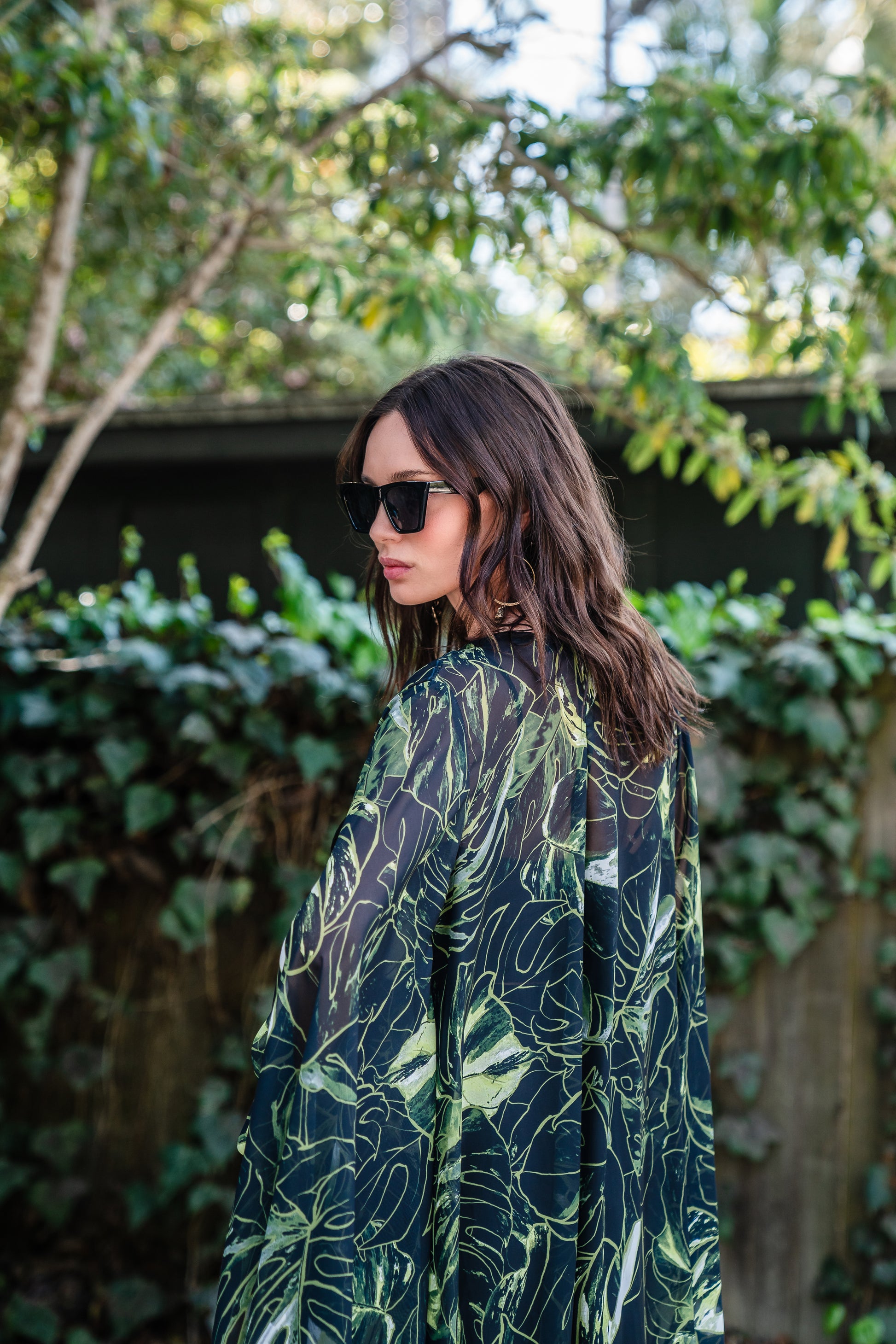 Model wears black chiffon Kimono with varying green palm leaf print. The kimono has classic wide kimono sleeves and lands at midi length. Kimono has a matching tie belt.