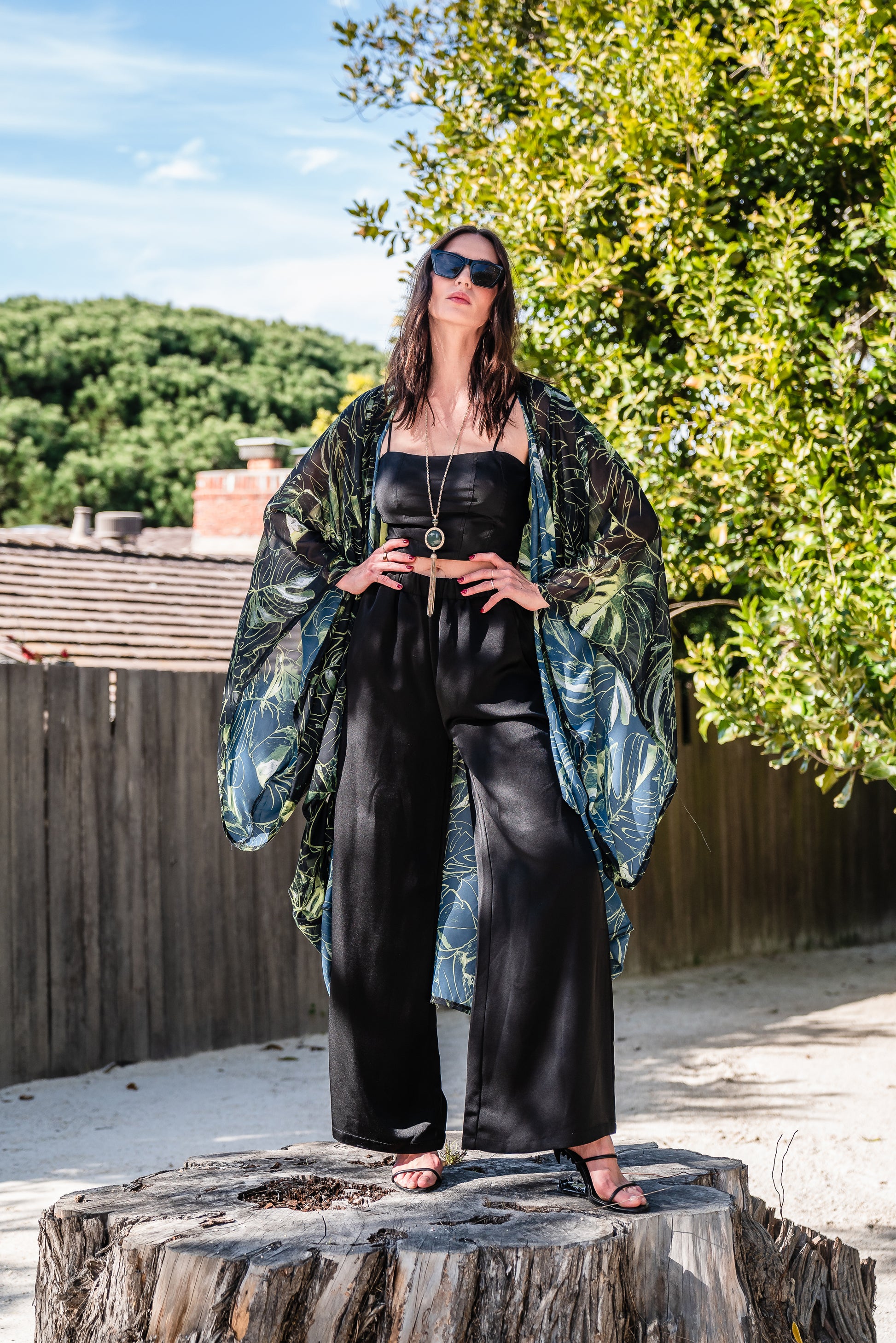 Model wears black chiffon Kimono with varying green palm leaf print. The kimono has classic wide kimono sleeves and lands at midi length. Kimono has a matching tie belt.
