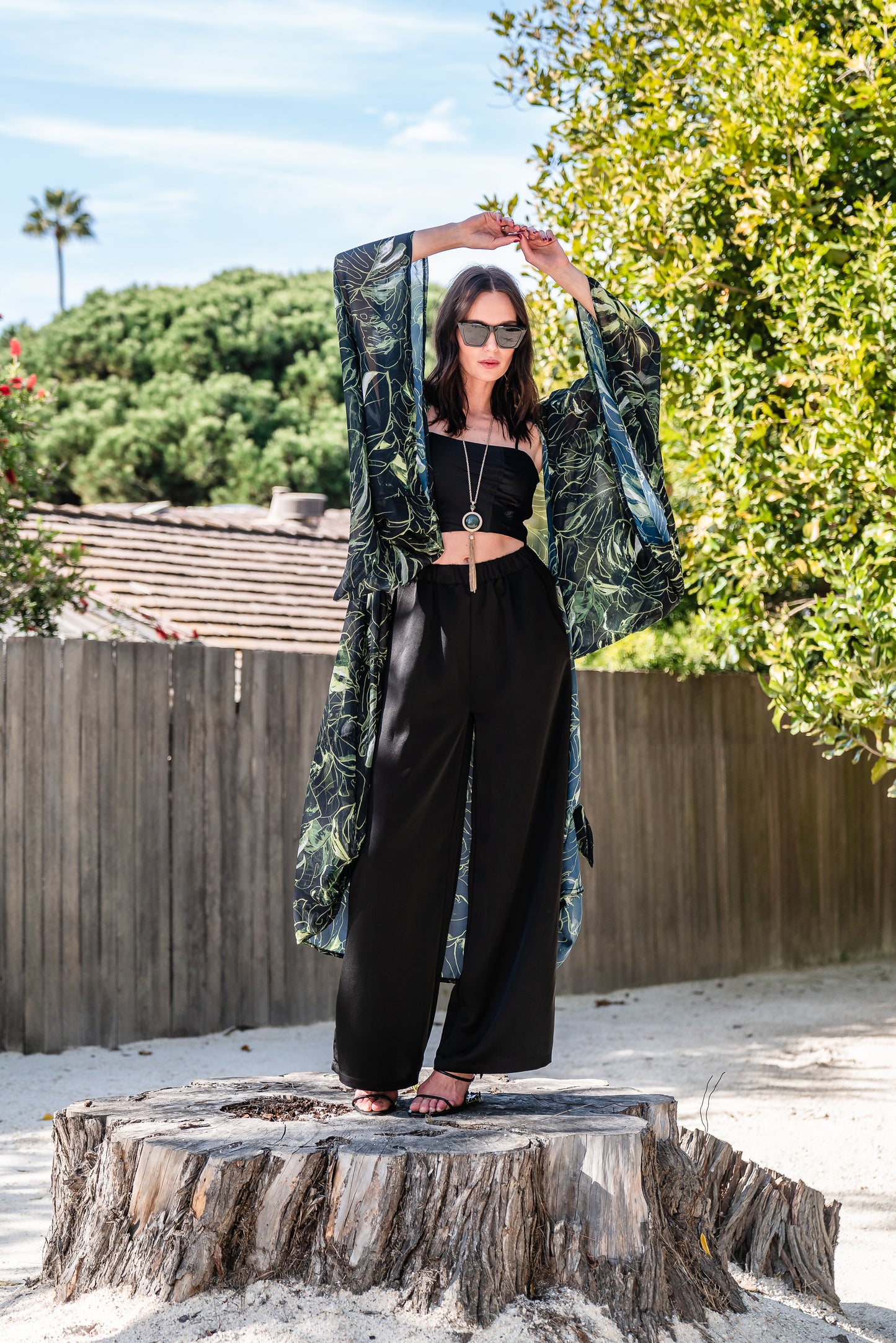 Model wears black chiffon Kimono with varying green palm leaf print. The kimono has classic wide kimono sleeves and lands at midi length. Kimono has a matching tie belt.