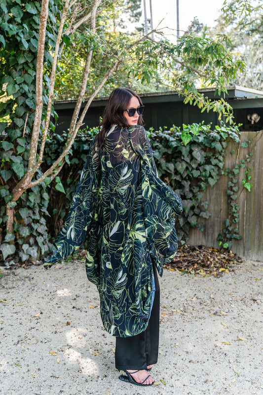 Model wears black chiffon Kimono with varying green palm leaf print. The kimono has classic wide kimono sleeves and lands at midi length. Kimono has a matching tie belt.