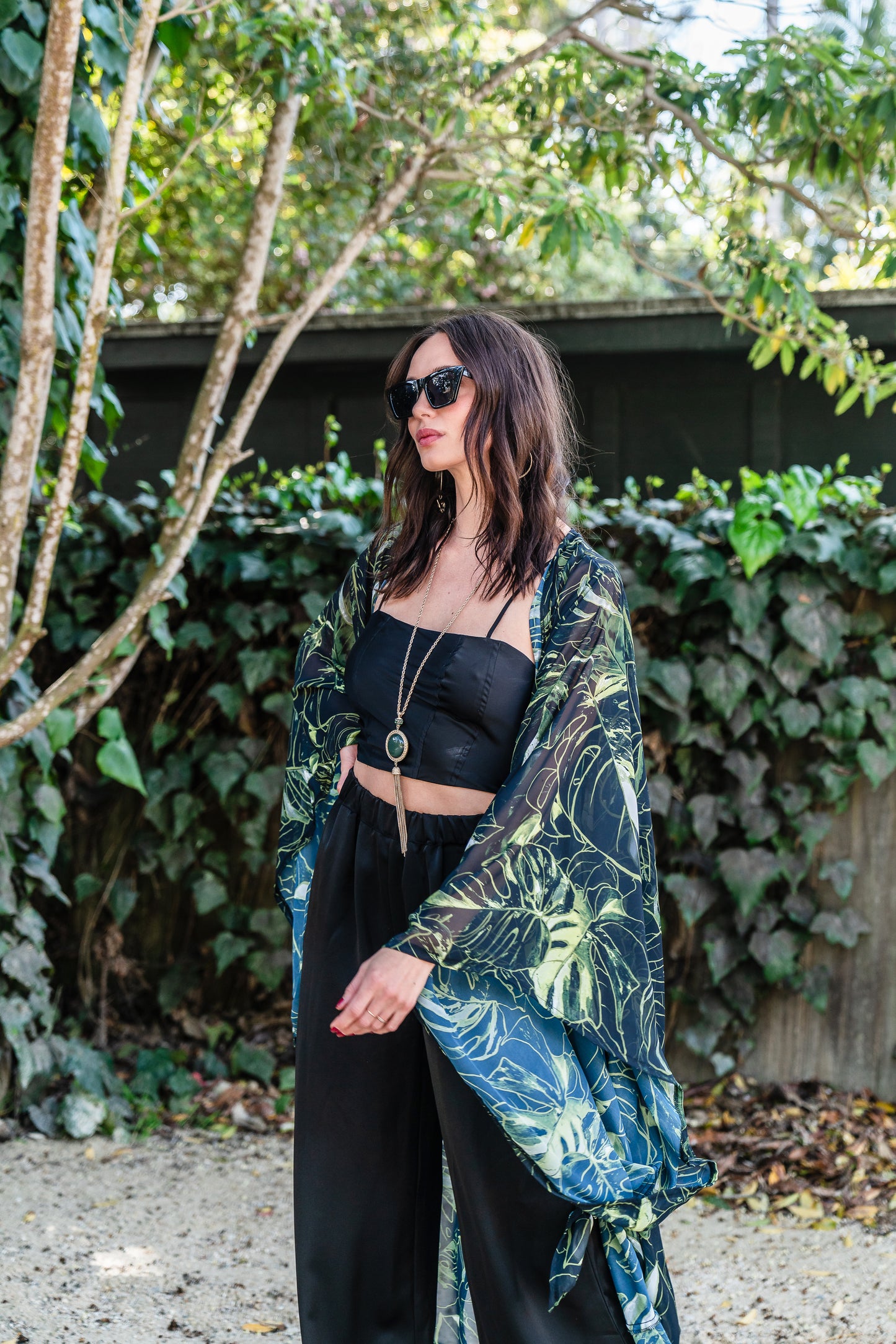 Model wears black chiffon Kimono with varying green palm leaf print. The kimono has classic wide kimono sleeves and lands at midi length. Kimono has a matching tie belt.